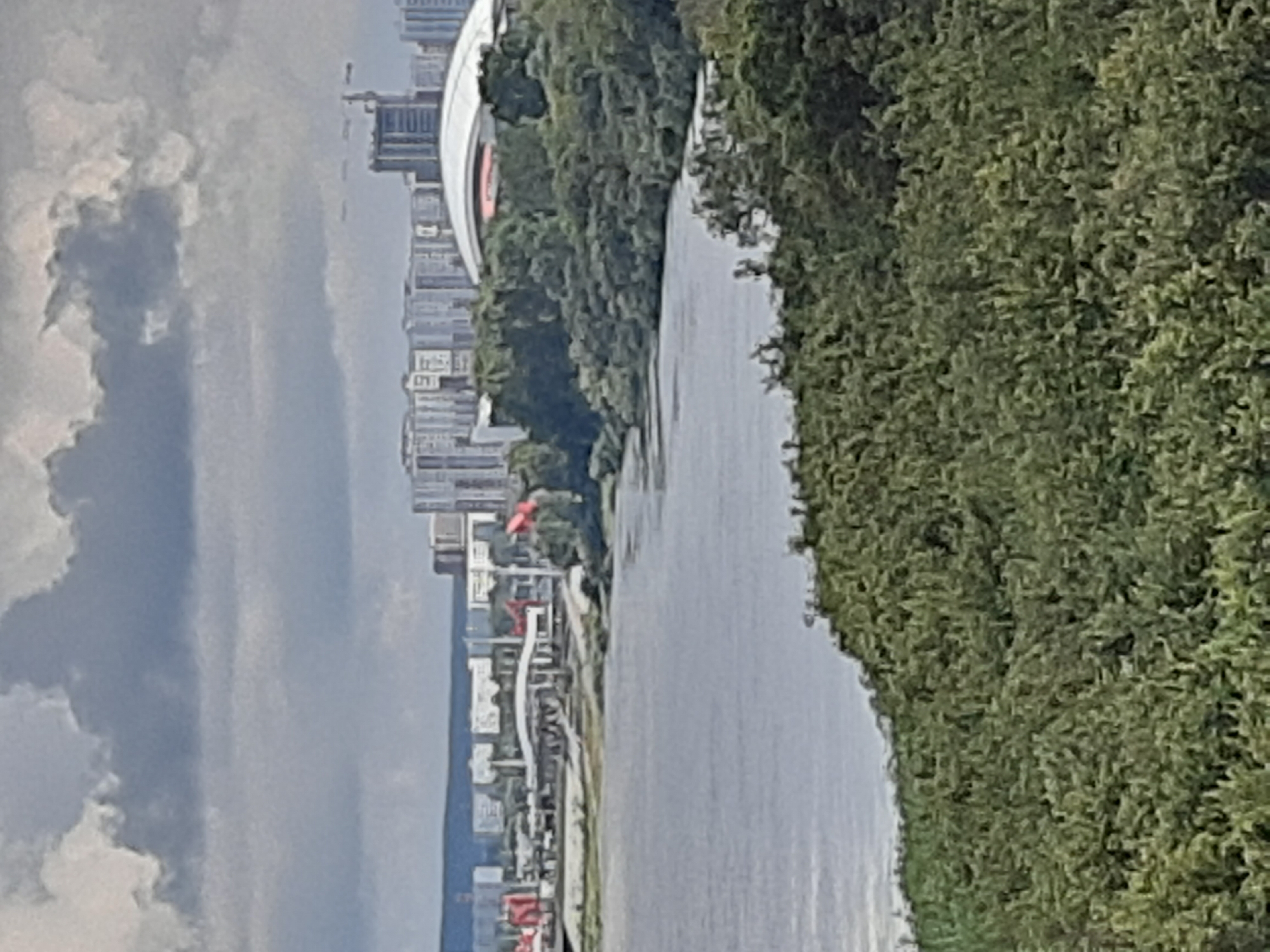 This is an outdoor, medium-distance, eye-level photograph taken on a cloudy day. In the foreground, a dense, green tree line runs along the bottom of the frame and extends up the right side, obscuring much of the lower portion of the scene. Beyond the trees, a wide, calm body of water fills the middle ground, reflecting the overcast sky. In the background, a cityscape is visible, featuring a cluster of modern high-rise buildings and a large, white, domed structure, likely a stadium or arena, with a red accent. Construction cranes are also present, suggesting ongoing development. The water in the foreground is the Tom River, and the cityscape is the city of Kemerovo, Russia.