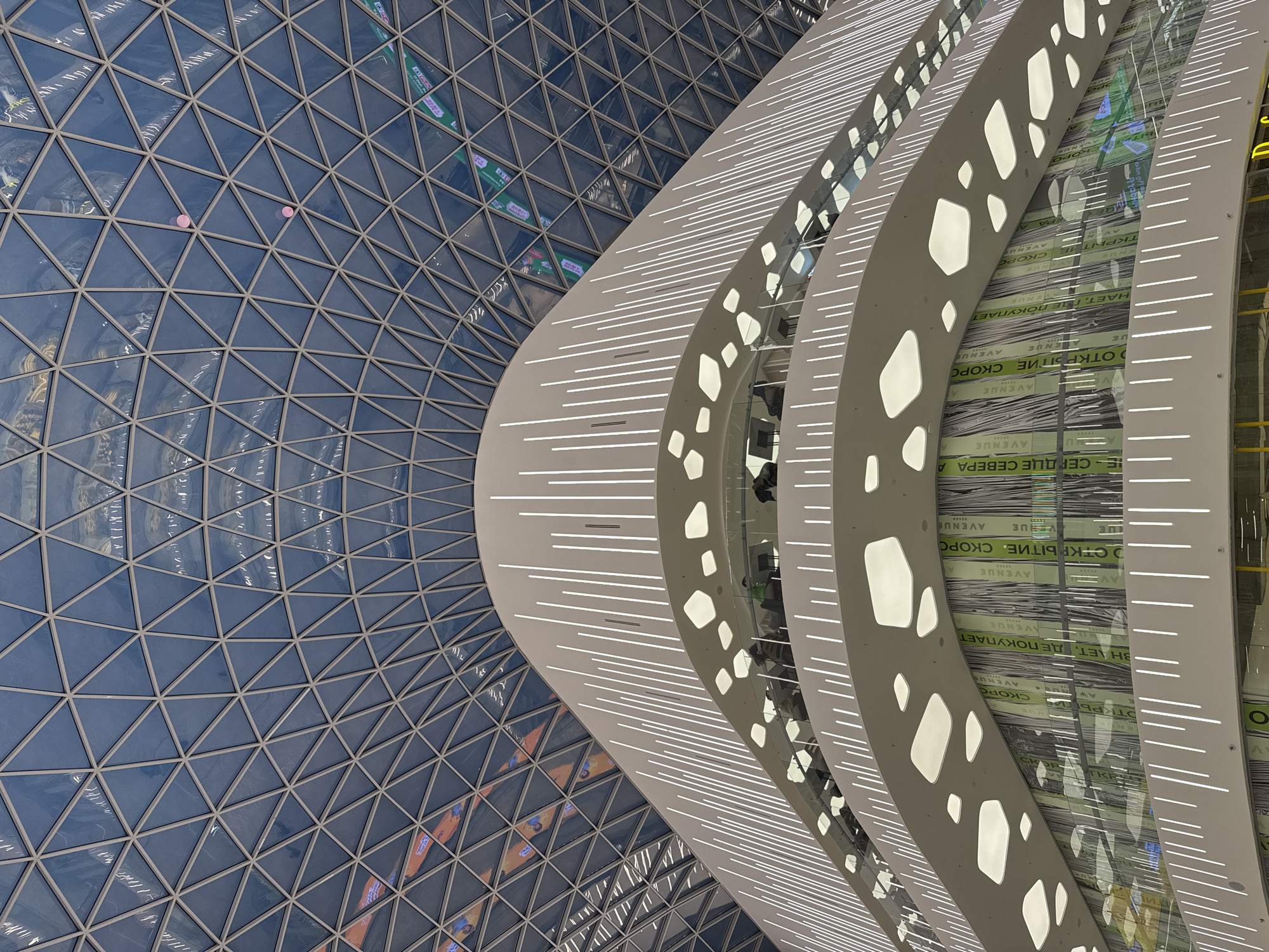 The image is an angled, upward view of an interior architectural design, specifically the Aviapark shopping center in Moscow, Russia. A triangular glass-paneled structure dominates the left side of the image, creating a geometric pattern that fades into the background. To the right, a multi-tiered, curved structure in a light beige color is prominent, featuring horizontal lines of light and irregular, rounded cutouts. The cutouts offer glimpses into the levels beyond, where text in Cyrillic and "AVENUE" can be seen. The architectural design has a modern, futuristic aesthetic with a focus on light and pattern.