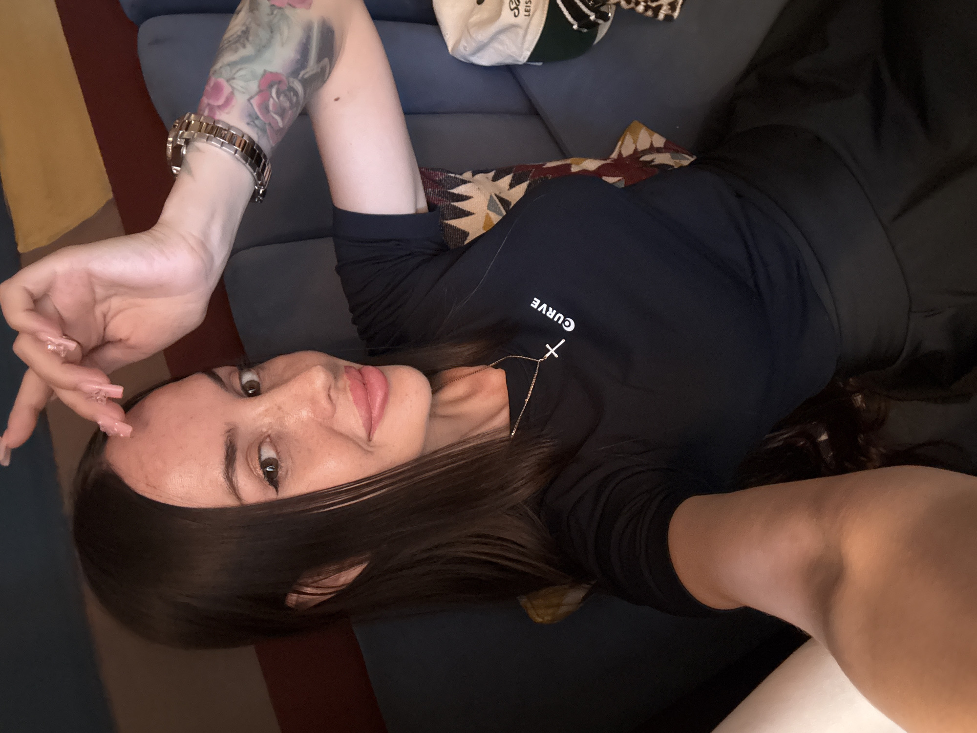 A woman is taking a selfie while sitting on a blue couch. She has long, dark brown hair and is wearing a black t-shirt with a small cross and the word "CURVE" on it, and a black skirt. She is wearing makeup, including eyeliner and lipstick. She has a watch on her left wrist and a tattoo on her left arm. A patterned pillow is visible behind her. A white and green baseball cap is on the couch to her right. The background shows a wall with horizontal stripes of red and yellow. The image does not provide any information about the location in Saint Petersburg, Russia.