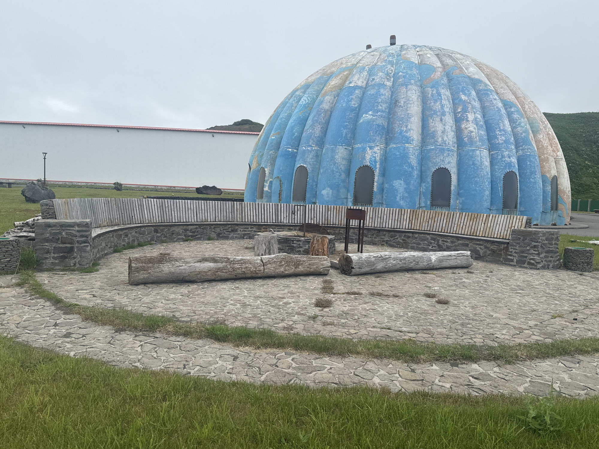 The image depicts an outdoor area with a large, blue and white geodesic dome structure in the background. The dome appears weathered, with peeling paint and visible seams. In front of the dome is a circular paved area with a stone wall and wooden seating. Several large logs are placed on the paved area, likely serving as seating or decorative elements. A metal barbecue grill is also present. The surrounding environment includes a white building with a red roof, green grassy areas, and rolling hills in the distance under an overcast sky. The scene is devoid of people. This area is located in Kuril'sk, Russia.