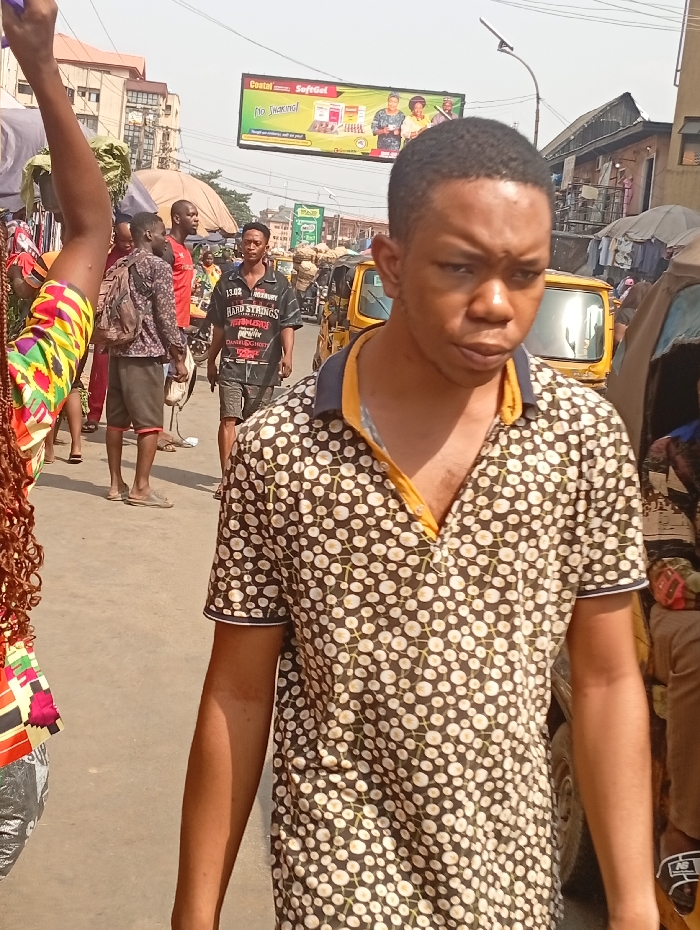 The image depicts a bustling street scene in Onitsha, Nigeria, under bright daylight. In the immediate foreground, a young adult male with dark skin and short dark hair is captured from the chest up, walking slightly to the viewer's left. He wears a dark polo shirt patterned with numerous small, light-colored circles connected by thin lines, featuring a yellow inner collar and a dark collar with a white stripe. His expression is neutral, with a slight furrow in his brow.
To his immediate left, partially visible, is the shoulder and arm of another individual, likely female, with long, reddish-brown curly hair and a colorful top featuring red, yellow, black, and green geometric patterns.
In the midground, several other individuals are present. One male, wearing a dark t-shirt with white text including "HARD STRINGS," "NEWBURY," and "DANIEL GHOST," and dark shorts, walks towards the viewer's right. Behind him, to the left, two males are conversing; one wears a dark patterned shirt and a backpack, while the other wears a red t-shirt. Multiple yellow three-wheeled vehicles, consistent with "keke Napep" tricycles, are visible along the street, parked or in motion, indicating public transportation.
The background features multi-story buildings, suggesting a commercial or residential urban area, with some appearing to house shops or stalls. Overhead, a large green billboard advertises "Coastal SoftGel" and states "No Smoking!", displaying images of three women. The street surface appears paved, likely concrete or asphalt. The overall scene is characterized by active pedestrian traffic and urban infrastructure typical of a busy Nigerian city street or market.