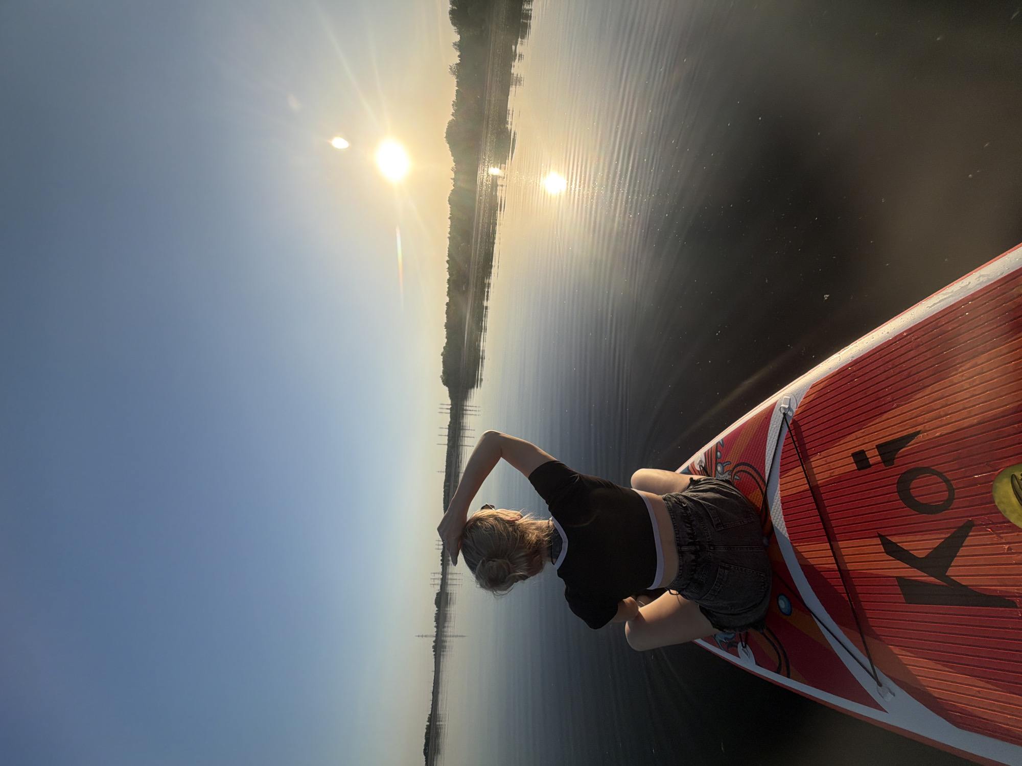 A high-angle shot shows a person sitting on a red and white stand-up paddleboard (SUP) on a body of water, likely a lake or wide river. The person is wearing a black crop top and dark denim shorts. Their back is facing the camera, and their left arm is raised, resting on their head. The board is positioned at the bottom right of the frame.
The water is dark and reflective, mirroring the bright sky. The sun is visible as a bright light source, reflecting off the water's surface. In the background, there's a strip of land with trees and what appears to be power lines or industrial structures in the distance. The sky is a gradient of blue to yellow, with the sun creating a hazy effect.
Based on the information provided, the image was taken in the city of Chaikovsky, Russia.