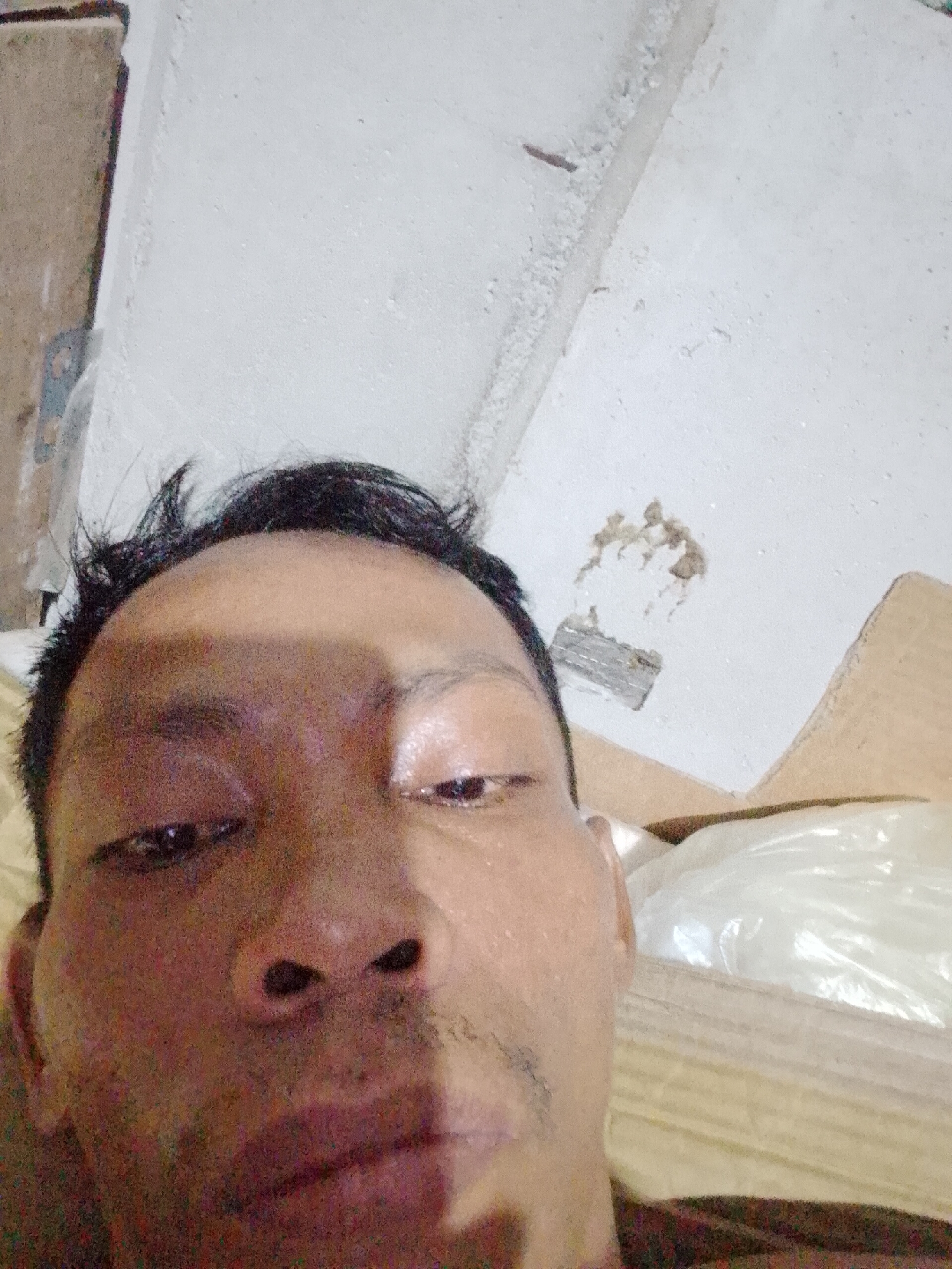 A close-up selfie of a man with dark hair and brown skin. The background is a textured white wall with damage and a cardboard box filled with a white plastic-wrapped object. The photo appears to have been taken indoors. No information regarding the location of Bukit Ketok, Indonesia can be discerned from this image.