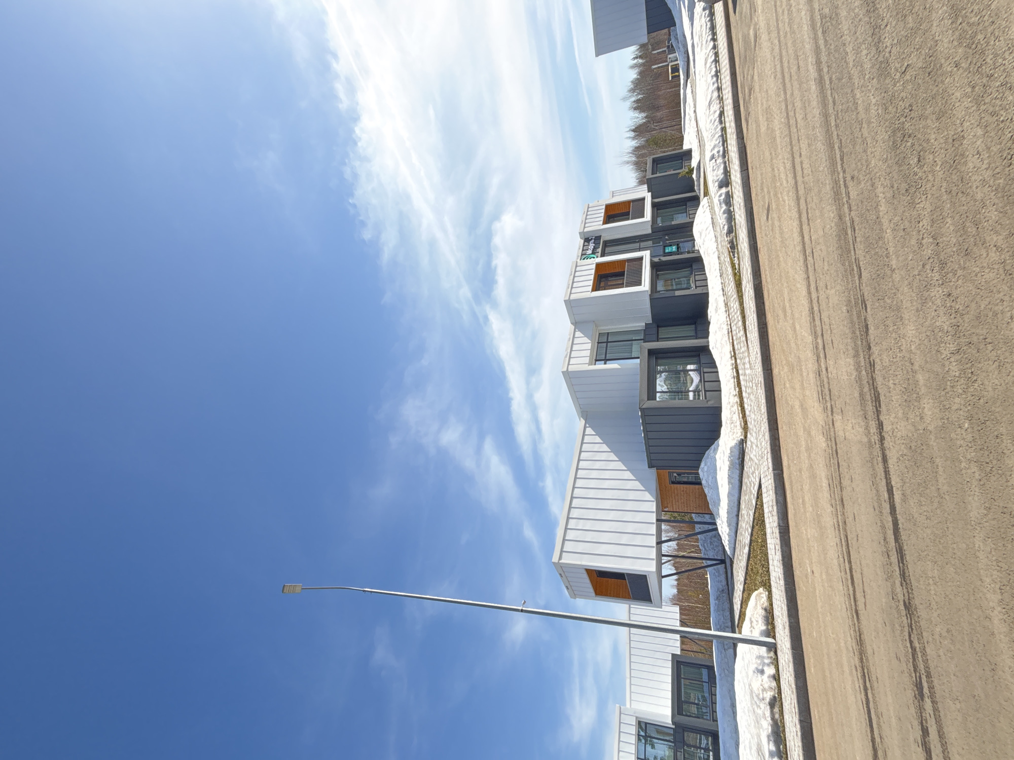 The image depicts a row of modern, boxy apartments situated along a paved street, likely in the city of Votkinsk, Russia. The apartments have a distinctive architectural style, featuring a combination of white and dark gray cladding with occasional orange accents around the window recesses. The architecture is clean and angular, and it appears as if the row of apartments extends into the distance.
The sky is a vibrant blue with thin, wispy cirrus clouds streaking across it. A tall, silver streetlamp is visible in the foreground against the sky. A narrow strip of land separates the apartments from the street, with patches of melting snow and a bit of green grass visible. The paved street is wide and devoid of traffic or pedestrians. The image is taken from a low angle, looking upwards towards the buildings and the sky. The perspective emphasizes the height and modern design of the apartments.