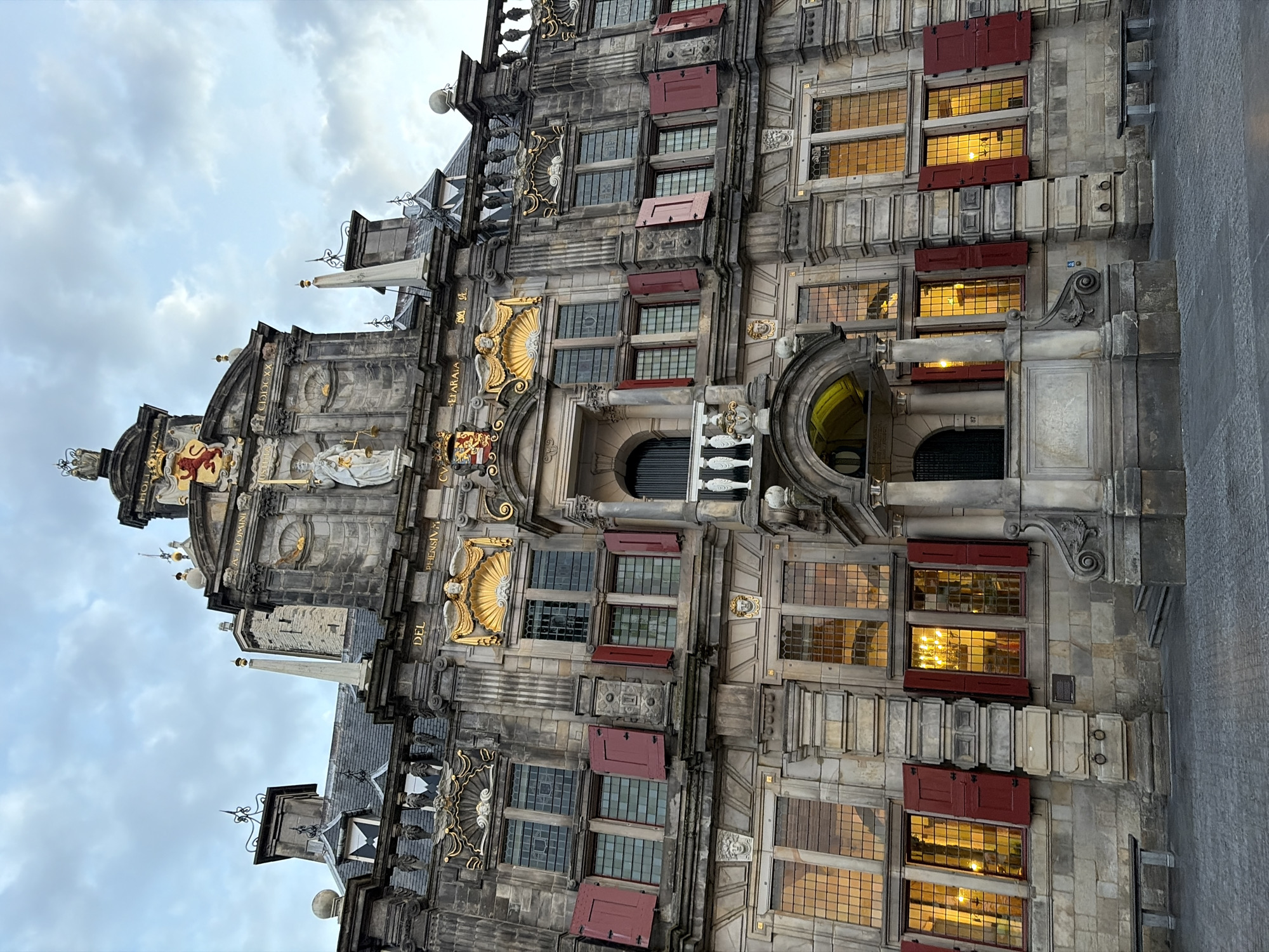 The image depicts the Stadhuis (City Hall) of Delft, Netherlands. The building is a multi-story structure of grey stone, with a highly ornamented facade in the Renaissance style. The windows are rectangular with multiple panes, some having red shutters that are partially open. The interior of the windows emit an orange light. The building features several sculptures and decorative elements, including gilded angels, coats of arms, and figures. The sky is overcast.