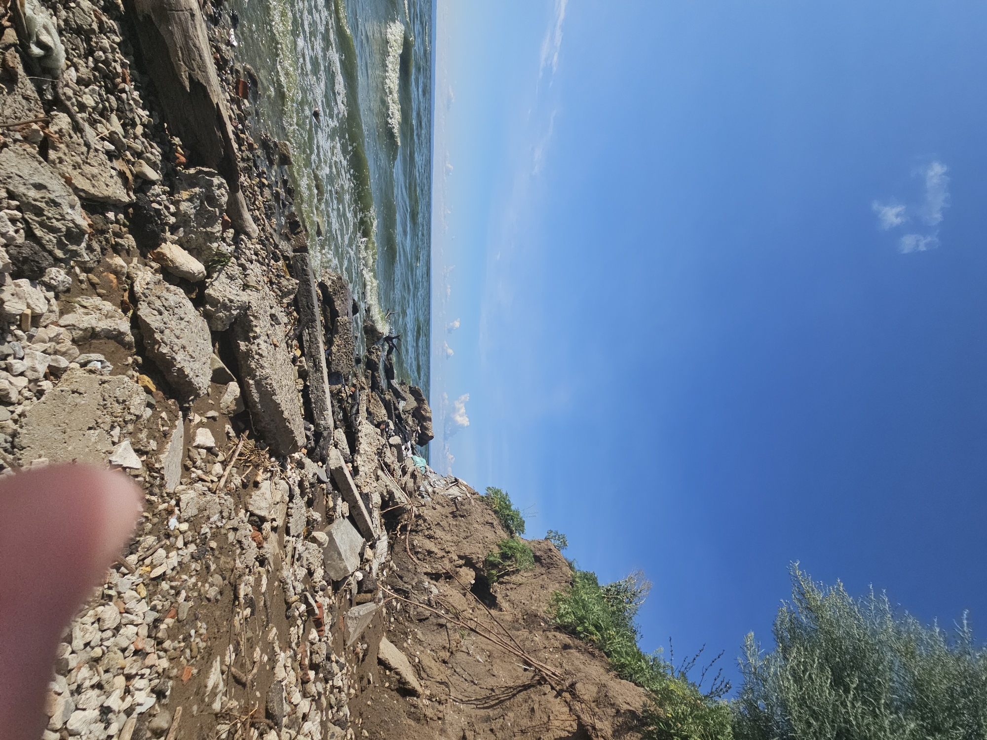 The image depicts a rocky shoreline with a steep, eroded cliff face on the right. The cliff is covered in vegetation and shows exposed soil and roots. Debris, including concrete slabs, bricks, and scattered rocks, litters the foreground and the base of the cliff. The sea, with choppy, green water, is visible on the left, meeting the shore with small waves and foam. The sky is a clear, bright blue with a few scattered white clouds. The scene appears to be a natural landscape altered by erosion and human debris.