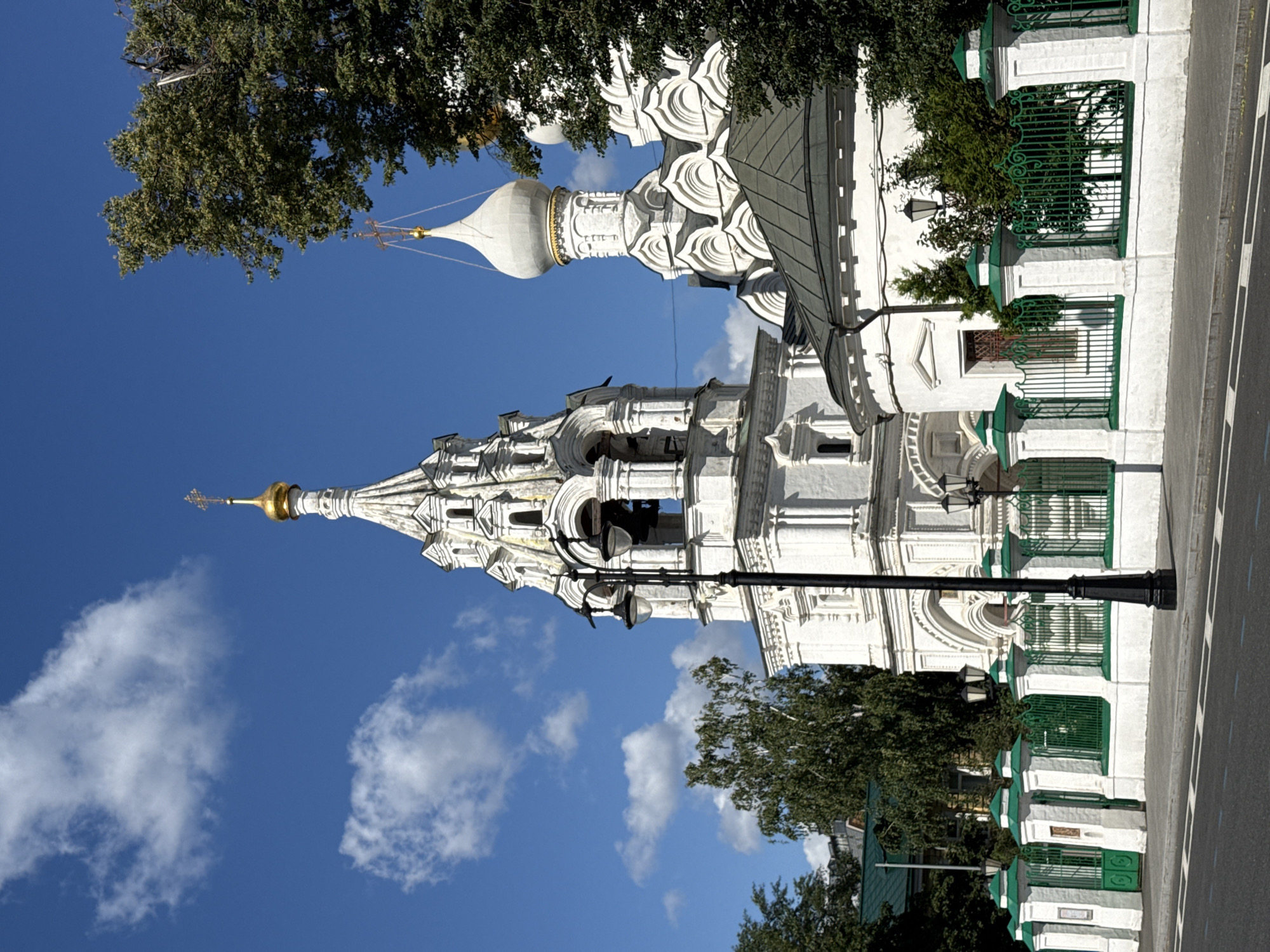 The image shows a white Orthodox church with a bell tower and a domed roof, located in Moscow, Russia. The church features golden crosses on its domes and detailed architectural elements. In front of the church is a white and green fence. A black lamppost with two lamps stands in the foreground, close to the fence. The sky is blue with white clouds, and trees with green foliage surround the building. The scene is captured during the daytime, with sunlight illuminating the structures.