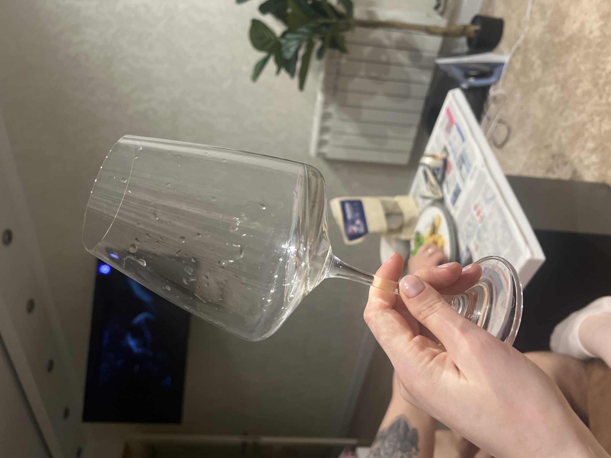 A person holds a wet, empty wine glass in their right hand. The person is likely sitting, and a portion of their leg is visible with a tattoo on the lower left. In the background are a television, a plant in a white planter, a white box, and a plate with food on it. The scene is lit with artificial lighting. Given the user's location, the image was potentially taken in Khimki, Russia.