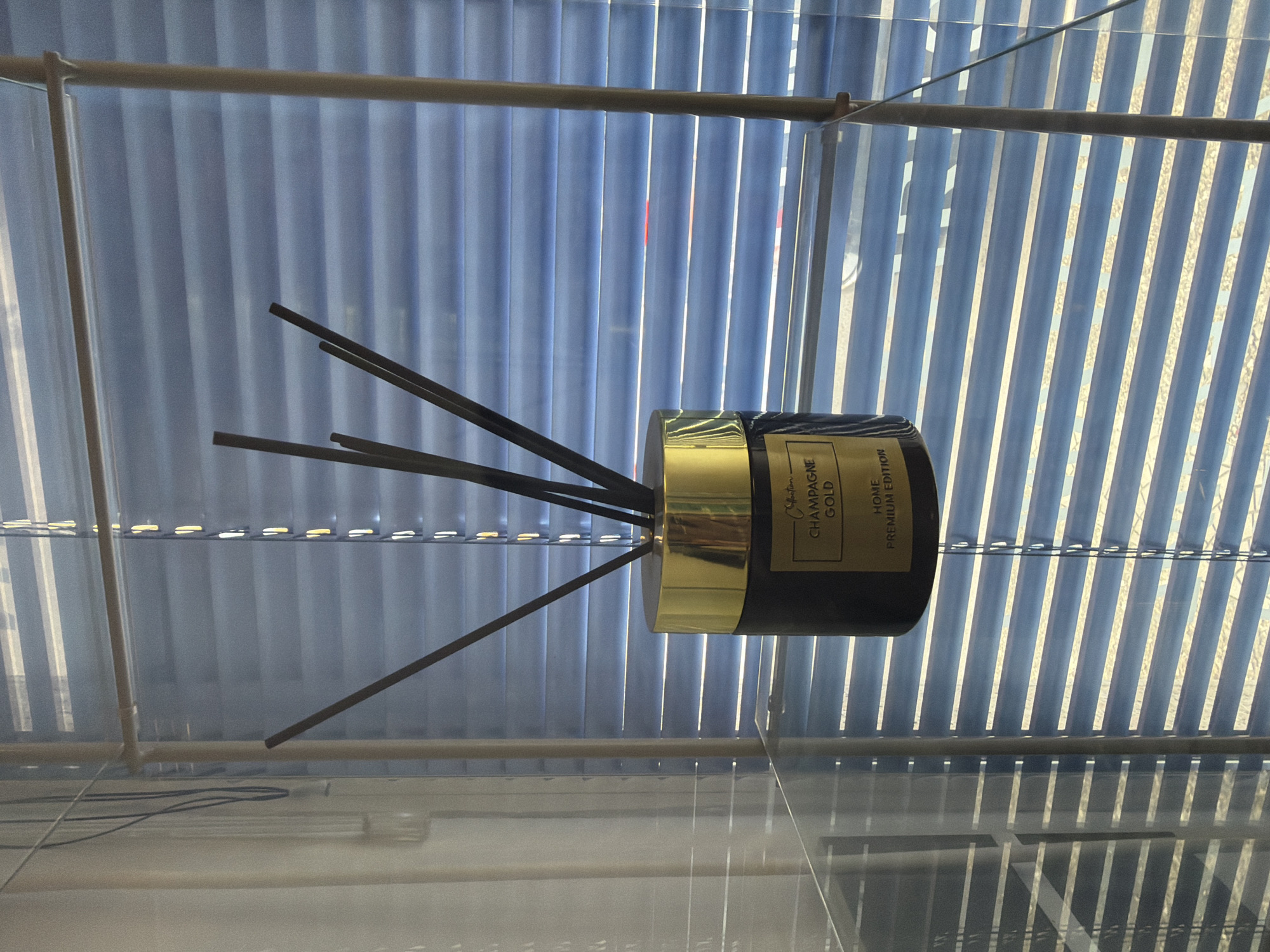 The image shows a reed diffuser with a gold lid and a black bottle with a gold label. The label reads "Collection CHAMPAGNE GOLD HOME PREMIUM EDITION." There are several dark reeds sticking out of the diffuser. The diffuser is placed on a glass shelf. In the background, there are blue blinds with sunlight coming through them. The scene appears to be indoors, likely in a home or store. The location is not specified in the image or the provided crops.