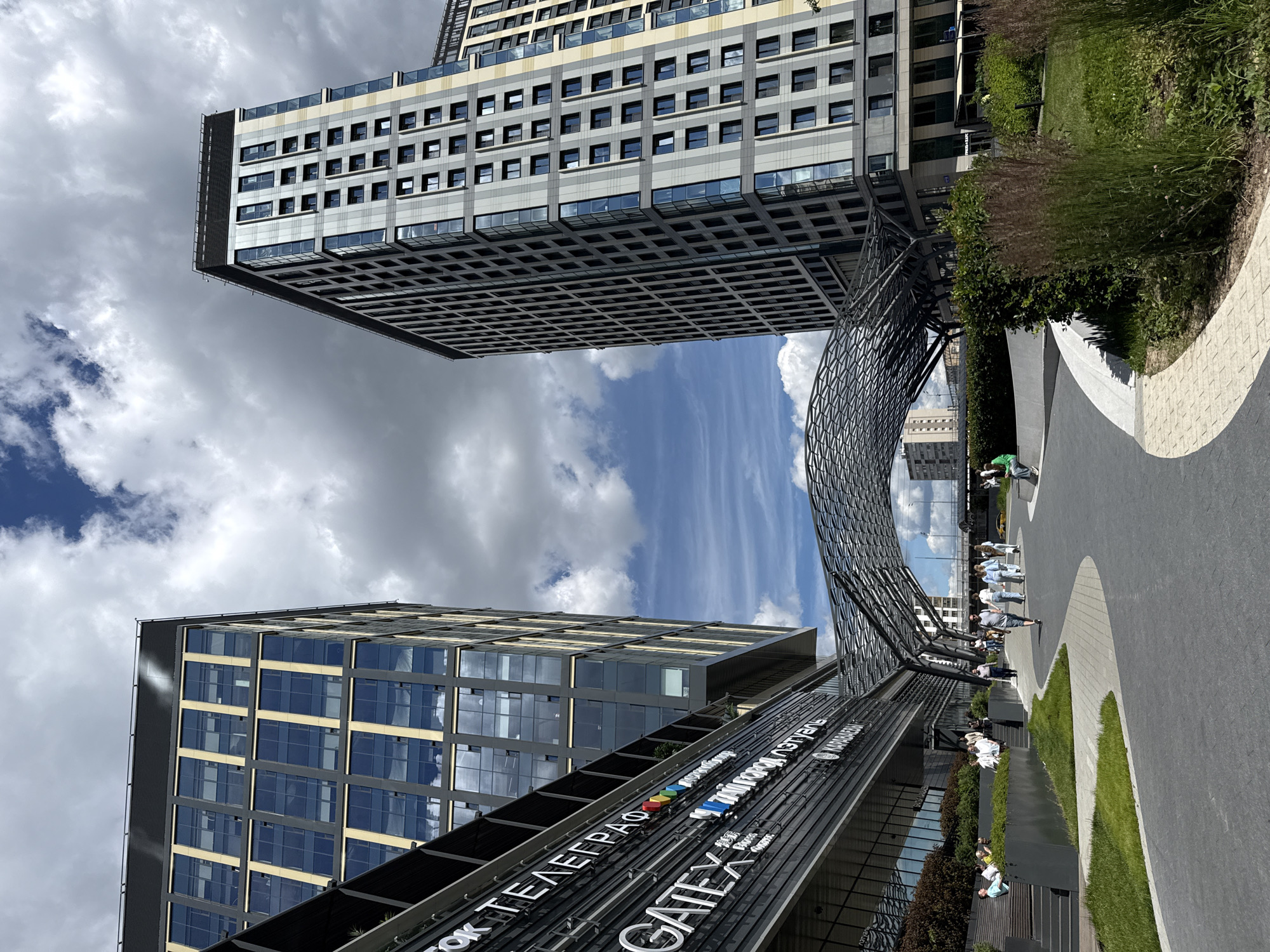 The image shows a modern urban scene in Moscow, Russia. Two prominent contemporary buildings with numerous windows dominate the frame, one of them extending outwards over a lower section. A large, curved metal structure, possibly a canopy or art installation, is positioned between the buildings.
In the foreground, a paved walkway curves through a landscaped area with grass and bushes. Several people are scattered throughout the scene, some walking and others sitting, suggesting a leisurely daytime activity. The sky is partly cloudy, with a mix of bright sunshine and overcast conditions, indicating pleasant weather. The time of day appears to be midday or early afternoon.
Visible text on a sign attached to the lower building reads "GATE" and "ТОК ТЕЛЕГРАФ". The overall mood is one of contemporary urban life and modern architecture.