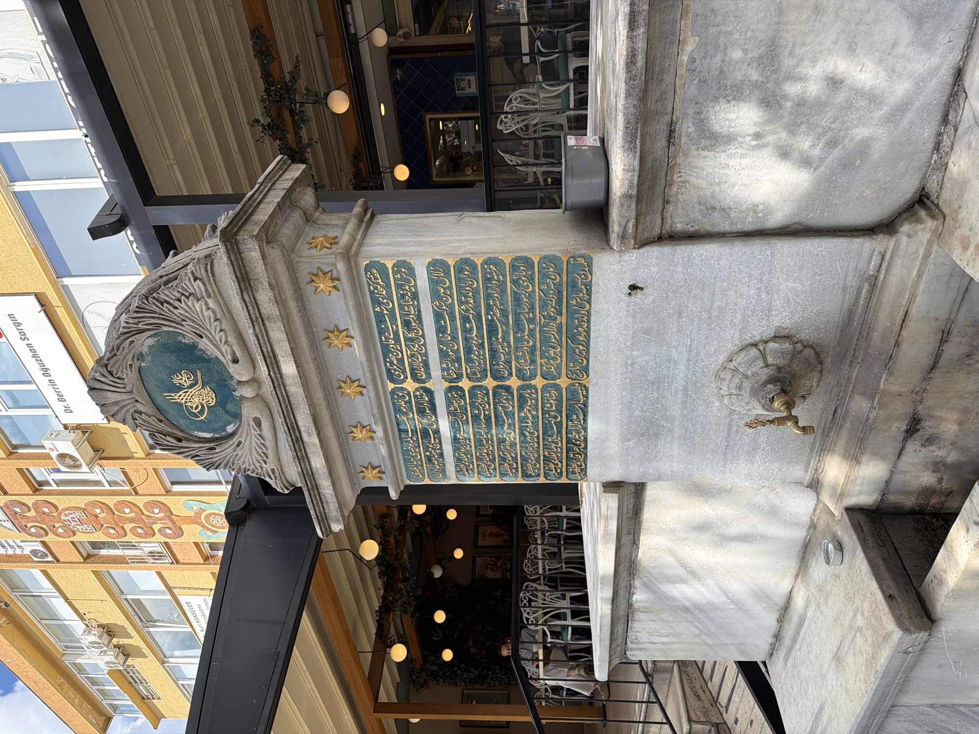 The image captures a street scene in Kadıköy, Turkey, focusing on a historic public fountain situated in front of a building. The fountain is constructed from marble and features intricate stone carvings, including a prominent emblem at the top. The central column of the fountain is adorned with a decorative plaque inscribed with ornate Arabic calligraphy, set against a teal background with gold accents. Below the calligraphy, a brass faucet is visible, indicating the fountain's functional purpose.
In the background, a yellow-colored building with multiple windows and air conditioning units suggests a commercial or residential structure. Signs in Turkish are partially visible on the building's facade. The area around the fountain appears to be an outdoor seating space for a cafe or restaurant, with tables and chairs set up under a retractable awning. Some patrons are seated at the tables, engaged in conversation or dining. Overhead lighting fixtures and greenery add to the ambiance of the establishment. The overall scene depicts a well-preserved urban landmark integrated into the daily life of the city.