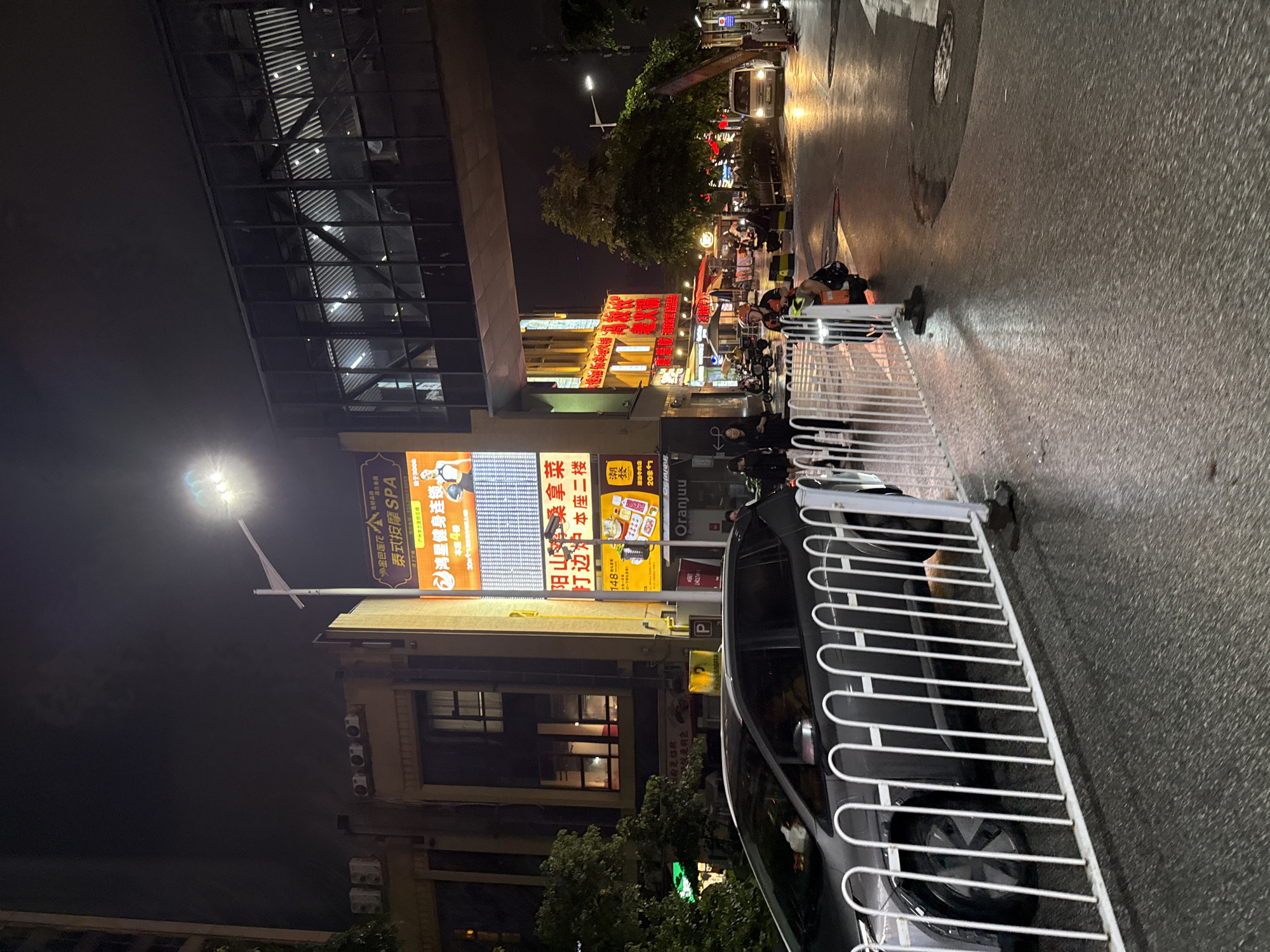 An evening street view in Haizhu District, China, shows a wet asphalt road reflecting streetlights and building lights. A white metal fence runs horizontally across the foreground, partially obscuring a black car on the left. Across the street, several businesses are illuminated, including a brightly lit building with a large yellow sign advertising "泰式按摩 SPA" (Thai Massage SPA) and another with prominent red characters, suggesting it's a restaurant. People are visible walking and riding scooters on the sidewalk and street. A white van is parked further down the road. An elevated pedestrian walkway, featuring a glass facade, spans across the street overhead. The overall atmosphere is that of a bustling urban environment at night.