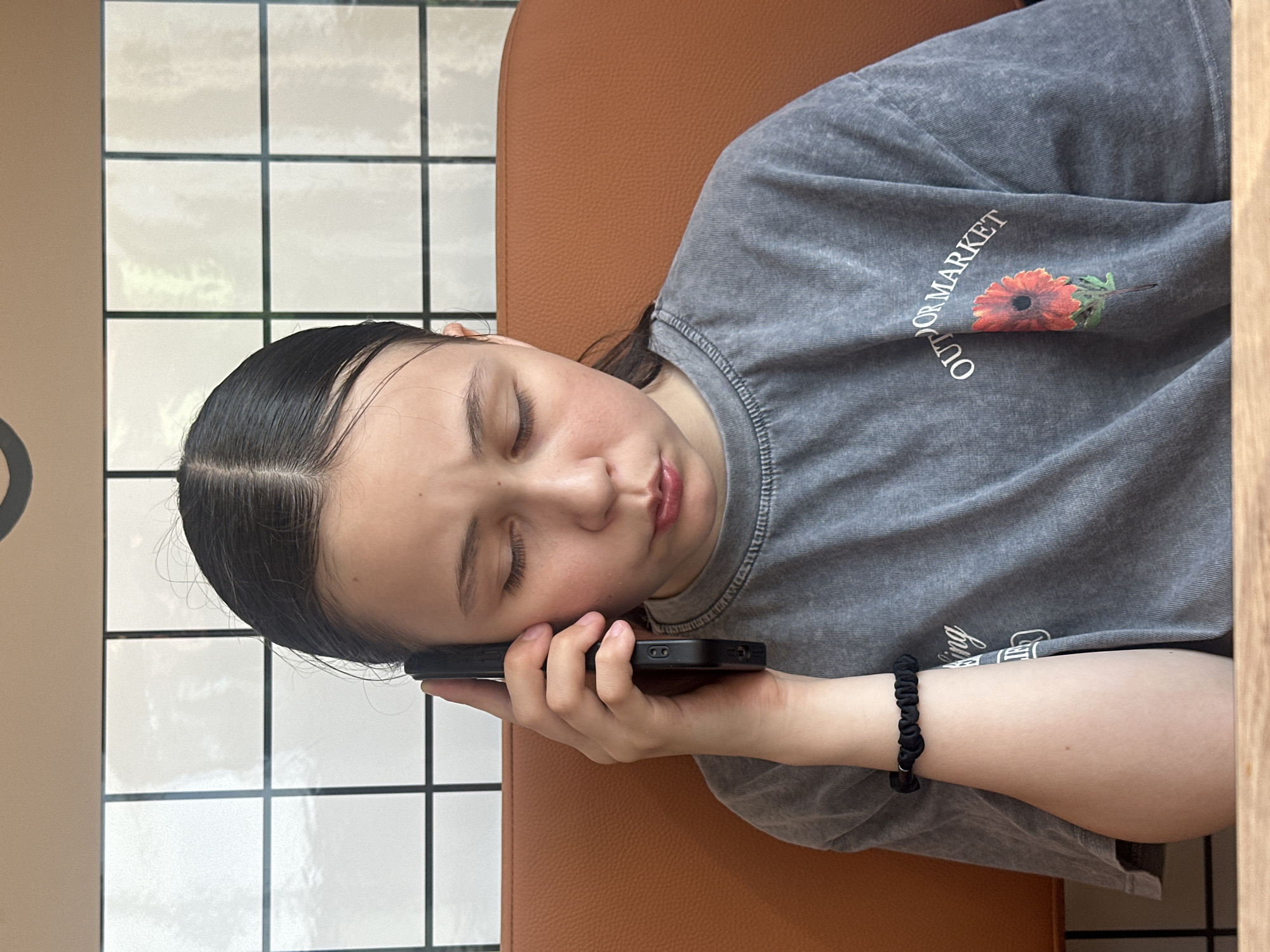 A young person rests their head on an orange chair with a black smartphone pressed to their ear. The person has fair skin, dark hair pulled back from their face, and is wearing a grey t-shirt with the word "OUTDOOR MARKET" and a floral graphic on the sleeve. The person wears a black scrunchie-type bracelet on their left wrist. The background includes a wall with square, white tiles and a wooden surface. There is no specific indication of this being Votkinsk, Russia in the image.