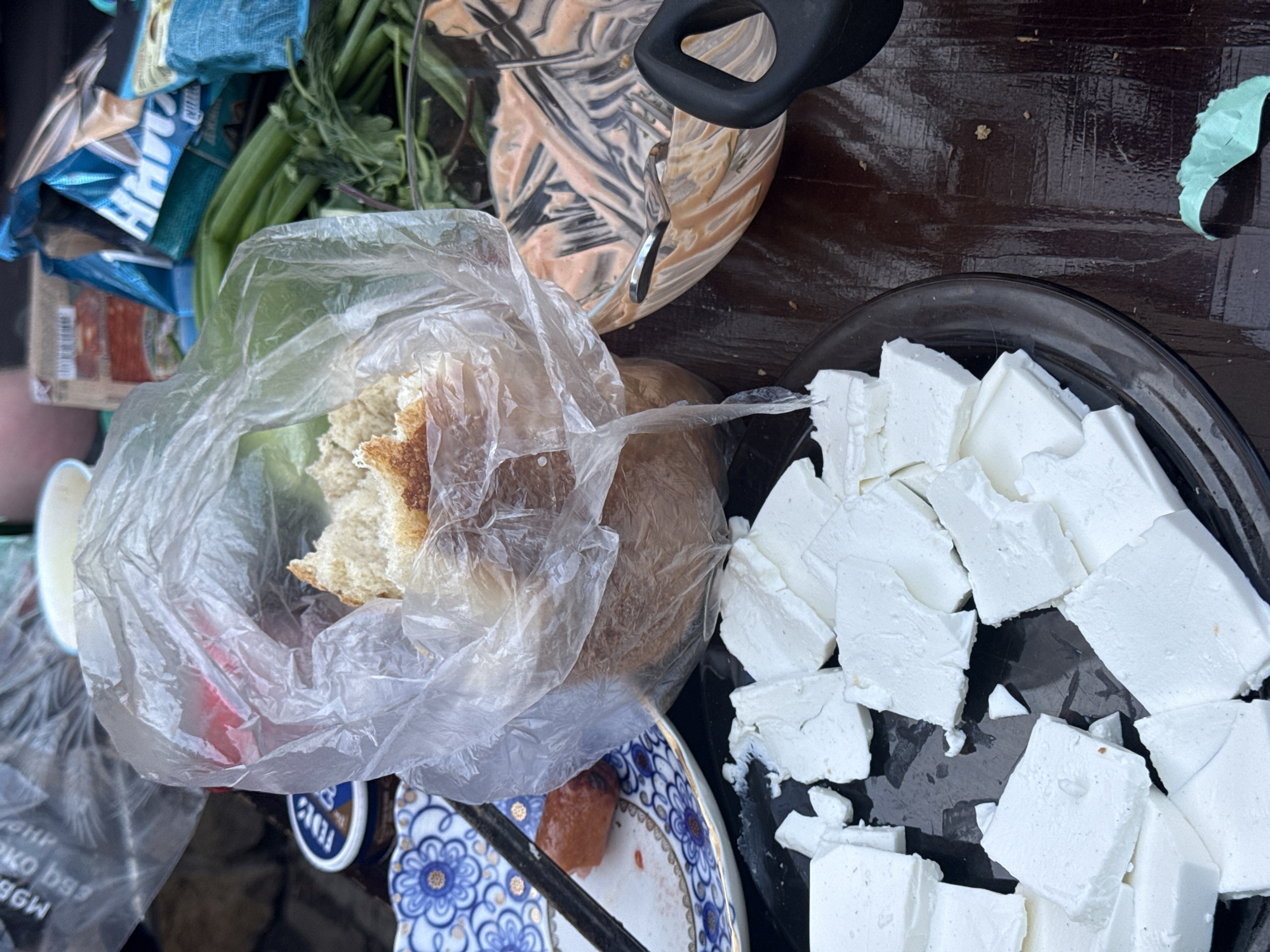 The image depicts a table in what appears to be an outdoor setting, possibly in Sukhobuzimskoye, Russia. A black plate is prominently displayed, filled with cubed white cheese. A plastic bag containing bread sits near the plate, alongside a bowl with a swirled tan and white mixture. Fresh green herbs or vegetables are present. A patterned plate with remnants of food is visible. Other items, including packaging, are also present on the table. The scene appears to be a casual meal setting.