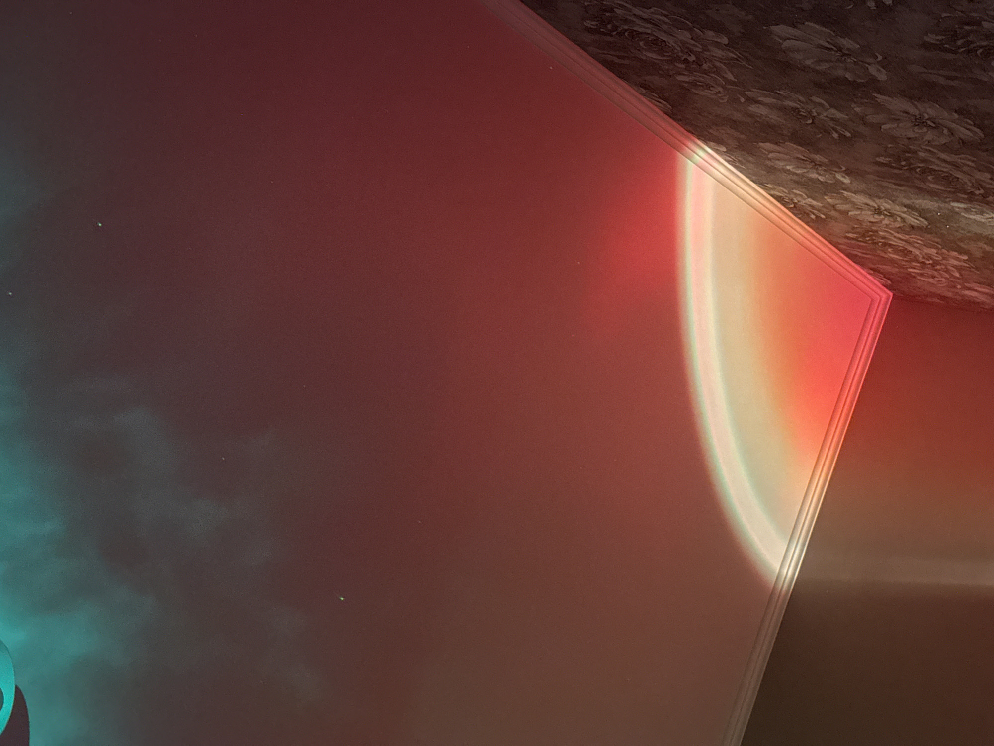 A projection of light, resembling a rainbow or sunset, casts vibrant hues of red, orange, and green onto a white ceiling and a floral-patterned wall. The light creates a gradient effect, with the brightest and most colorful part appearing to curve along the corner where the ceiling meets the wall. The ceiling is smooth and white, while the wall is covered in textured wallpaper with a dark, floral print. The scene is dimly lit, with the projected light being the primary source of illumination. The image is a close-up, focusing on the interplay of light and surfaces, with no people or distinct objects clearly visible besides the wall and ceiling. The location is described as Unknown, Unknown, suggesting an unspecified or generic indoor setting.