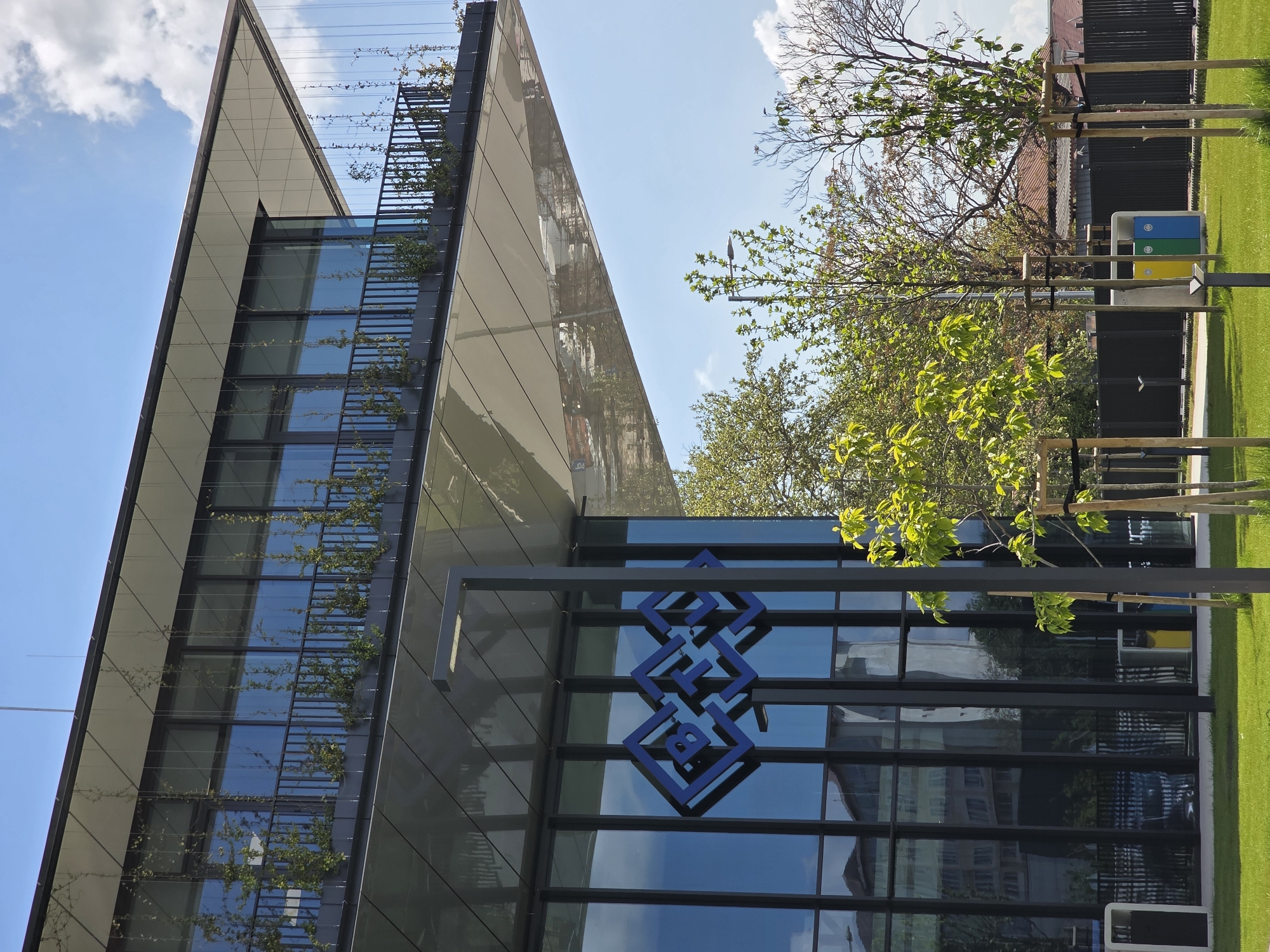 The image shows a modern building in Plovdiv, Bulgaria, possibly an office or commercial space. The building has a sleek design with a combination of glass windows and light-colored panels. Greenery is incorporated into the architecture, with plants growing along a trellis or support structure attached to the building's facade. The logo of the Bulgarian Development Bank (BDB) is prominently displayed on the building's glass exterior. The surrounding environment includes well-maintained green spaces with grass and young trees. A multi-colored recycling bin is visible, suggesting an emphasis on sustainability. The sky is partially visible with some clouds, indicating a sunny day.