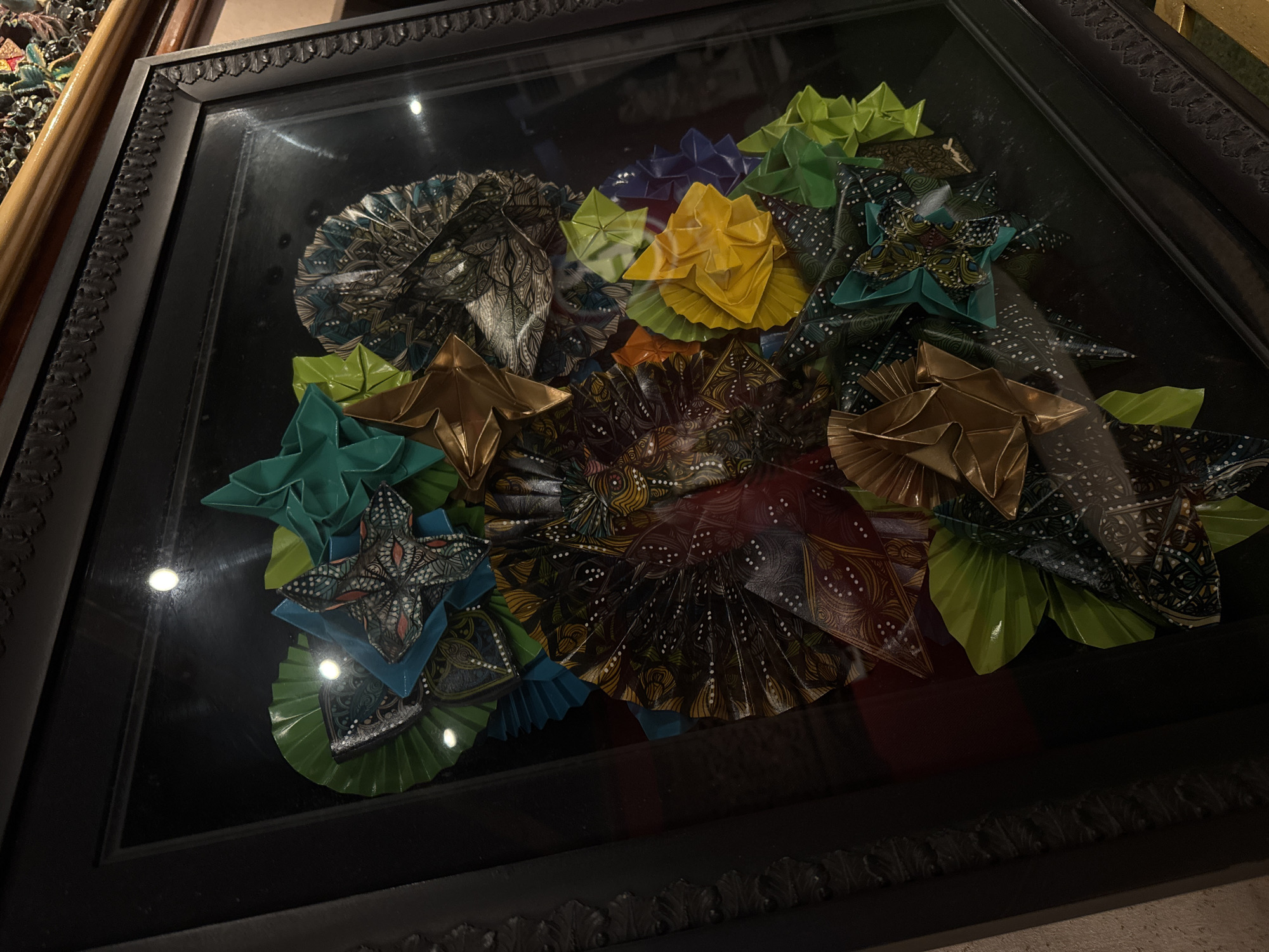 The image is a close-up, eye-level shot of a framed art piece consisting of folded paper flowers and decorative fans. The artwork is displayed against a dark background, possibly a wall. The frame is black with ornate detailing along its inner edge. The folded paper elements are arranged in a clustered, organic pattern, featuring a variety of colors including teal, green, yellow, purple, gold, and patterned fabrics in shades of brown, black, and white with intricate designs. Some pieces resemble fans, while others are star-shaped or petal-like. The lighting in the scene creates reflections on the glass covering the artwork, with several bright circular highlights visible. The overall impression is one of a handcrafted decorative item, possibly part of an interior design or a gallery display. No people are visible in the image. The location is Kuta, Indonesia.
