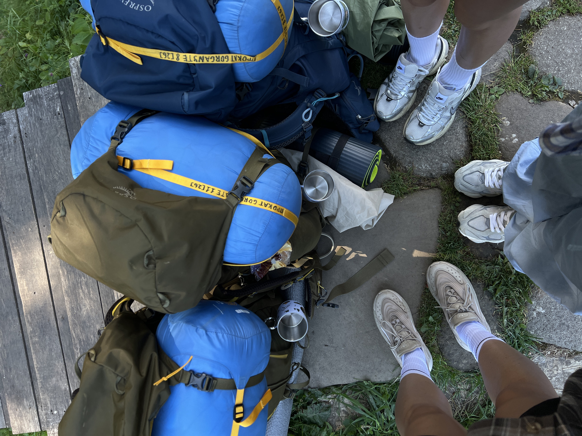 The image depicts a top-down view of several backpacks and camping gear arranged in a semi-circle on a wooden walkway and surrounding stone and grass. The backpacks are a mix of olive green and bright blue, with yellow straps and some visible branding. Attached to some of the blue bags are rolled sleeping bags. There are also two metal camping mugs and a rolled-up sleeping pad. In the foreground, parts of four individuals' legs and feet are visible, wearing athletic shoes and socks. The setting appears to be outdoors, with grass and foliage visible on the periphery. The image does not contain any explicit information or visual cues that definitively place the scene in the city of Vorokhta, Ukraine, or any other specific urban location.