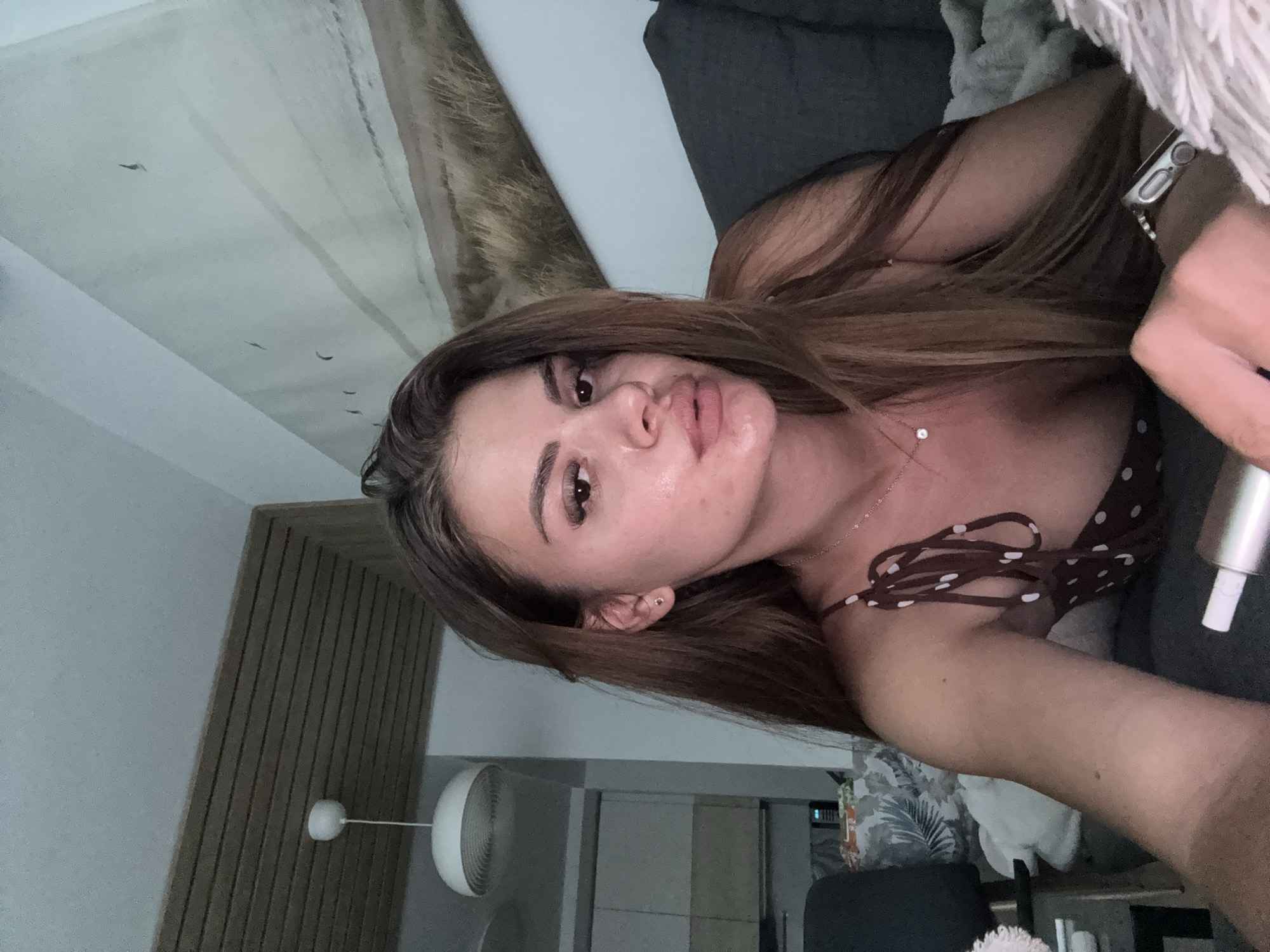 The image shows a young woman lying on a couch in what appears to be an indoor setting. She has long, brown hair and is wearing a brown and white polka dot top. She is looking towards the camera with her mouth slightly open. The background includes a wall with a landscape painting, a ceiling with wooden accents and a light fixture, and a dark gray couch. A small silver earring is visible on her left ear. She is wearing a necklace with a single clear pendant. On her left wrist, she has a smartwatch. Her right hand is holding a silver object that appears to be a vape pen. The lighting in the room is dim. There is no indication of the location being in the city of Plovdiv, Bulgaria.