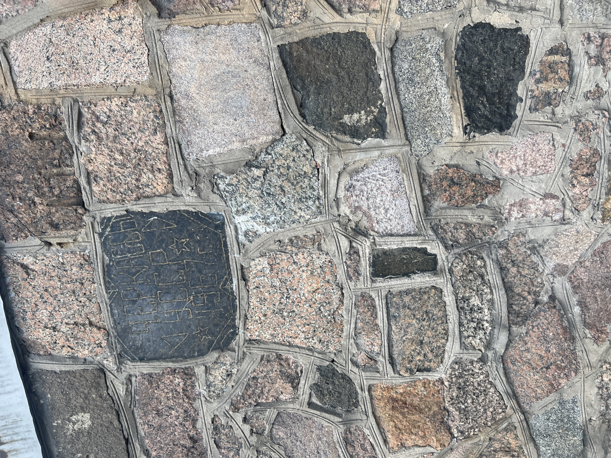 The image displays a close-up of a wall or paving surface made of irregularly shaped and sized stones set in light grey mortar. The scene depicts a textured architectural detail, likely from Saint Petersburg, Russia, given the granite materials common to the region.
The primary subjects are numerous stone blocks and the mortar binding them. Most stones are a type of granite, exhibiting speckled patterns in shades of pinkish-brown, light grey, dark grey, and black. Their edges are generally uneven, and they are arranged in a mosaic-like pattern. The mortar fills the gaps, and thin, incised lines run along the edges of many stones, creating a decorative or structured outline.
No specific activity or event is taking place; the image focuses on the static surface. The bright, even lighting suggests it is daytime, possibly overcast, as there are no harsh shadows.
A notable detail is a prominent dark, almost black, rectangular stone positioned in the bottom left-center. This stone features carved or etched inscriptions. Visible text and symbols include two five-pointed stars, one above and one below a central block of geometric patterns. The central block contains a series of highly stylized, angular symbols or characters that resemble an antique or unique script, possibly Cyrillic but heavily abstracted, or even runic. These symbols are arranged in horizontal rows, appearing to form words or a code, though they are not immediately legible as standard text. The overall design on this dark stone suggests a decorative or historical marker.