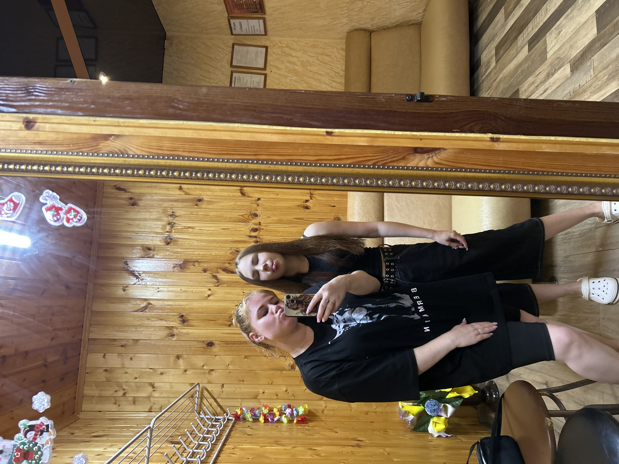 Two young women are taking a selfie in a room with wood-paneled walls. The woman on the left is wearing a black t-shirt and black shorts, and she is holding a phone to take the picture. She has her lips puckered. The woman on the right is wearing a black tank top and black pants. She is sticking her tongue out. In the background, there is a mirror, a sofa, and a shelf with decorative items. The room appears to be a private dwelling or a small establishment. There are no visible indicators of the specific location in Opary, Russia, beyond what can be inferred from the interior design.