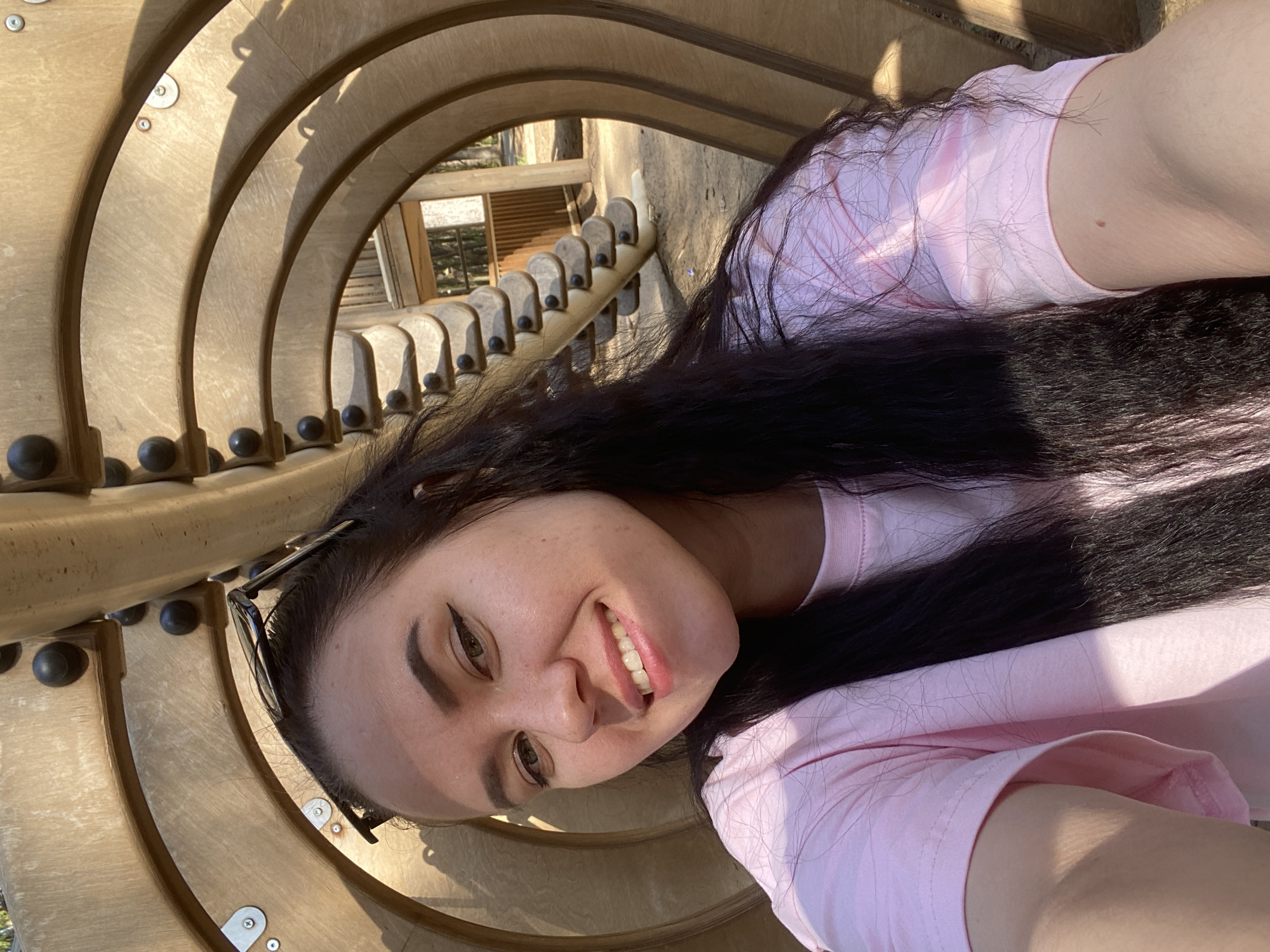 A woman is positioned inside a wooden tunnel structure, likely at a playground. She is fair-skinned, with long dark hair, and is wearing a light pink t-shirt. She has sunglasses pushed up on her head and is smiling toward the camera. The tunnel is composed of multiple curved wooden slats, supported by black knobs and metal fasteners. In the background, a building with windows is visible. The photo was taken in Perm, Russia.