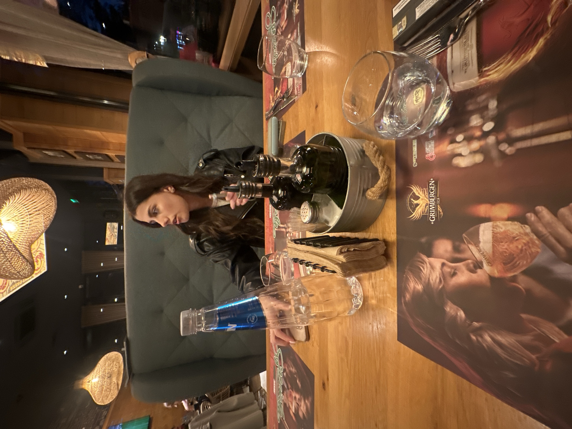 A woman with long brown hair sits at a wooden table in a restaurant. The table is set with a placemat that has an image of a person drinking beer. There's a metal bucket with wine bottles inside, a glass of water, a water bottle, and a stack of napkins. The restaurant setting is further indicated by the booth seating and decorative light fixtures. It is implied to be Plovdiv, Bulgaria, based on the name Grimbergen, a famous Belgian Beer which is popular in Plovdiv.