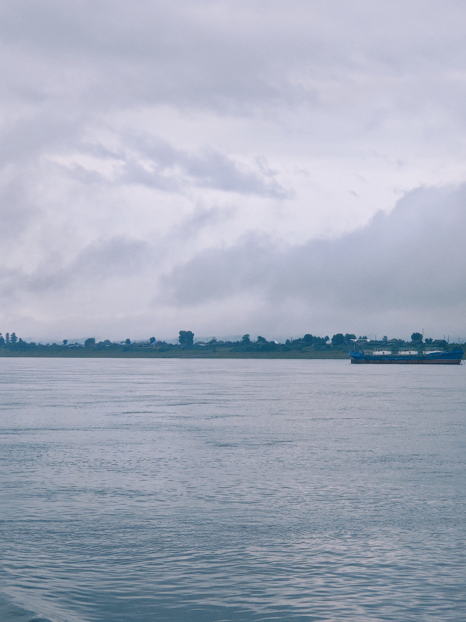 The image depicts a wide, calm river under an overcast sky. In the distance, a low-lying treeline with scattered buildings suggests a settlement on the opposite bank, possibly in Lesosibirsk, Russia, given the typical geography of Siberian river towns. A long, blue barge is present on the right side of the river, heading towards the left. The water's surface shows gentle ripples, reflecting the muted light of the cloudy day. The overall atmosphere is subdued and peaceful.