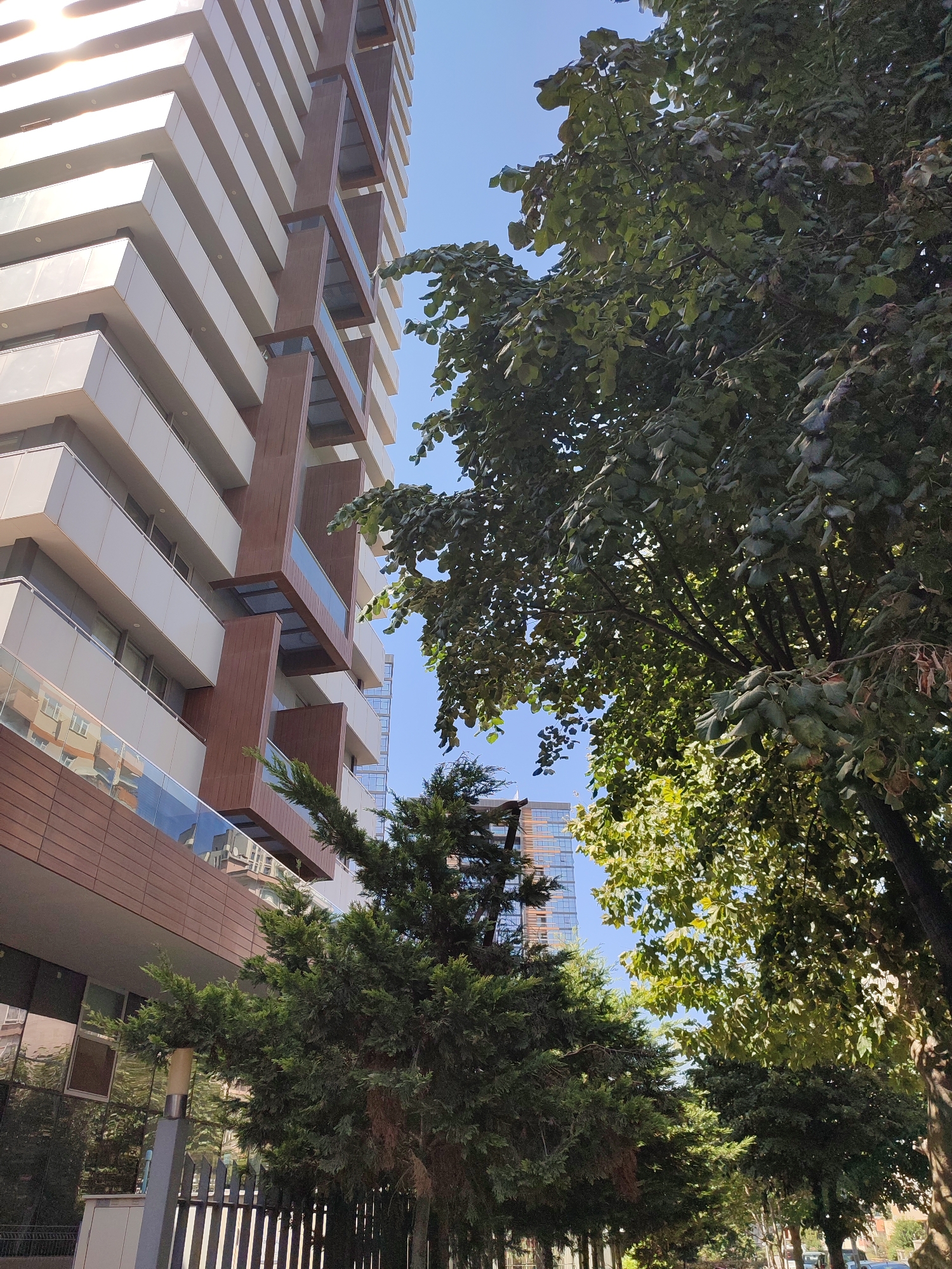 The image captures a view of a modern residential building in Kadıköy, Turkey, on a clear, sunny day. The building features a striking facade with alternating horizontal bands of white panels and darker brown wooden elements, punctuated by glass balconies. Tall, mature trees with dense green foliage partially obscure the building from the right and center. A clear blue sky serves as the background. In the foreground, a metal fence runs horizontally, bordering a sidewalk or street area where the base of the trees is visible. The scene appears to be an urban residential area.