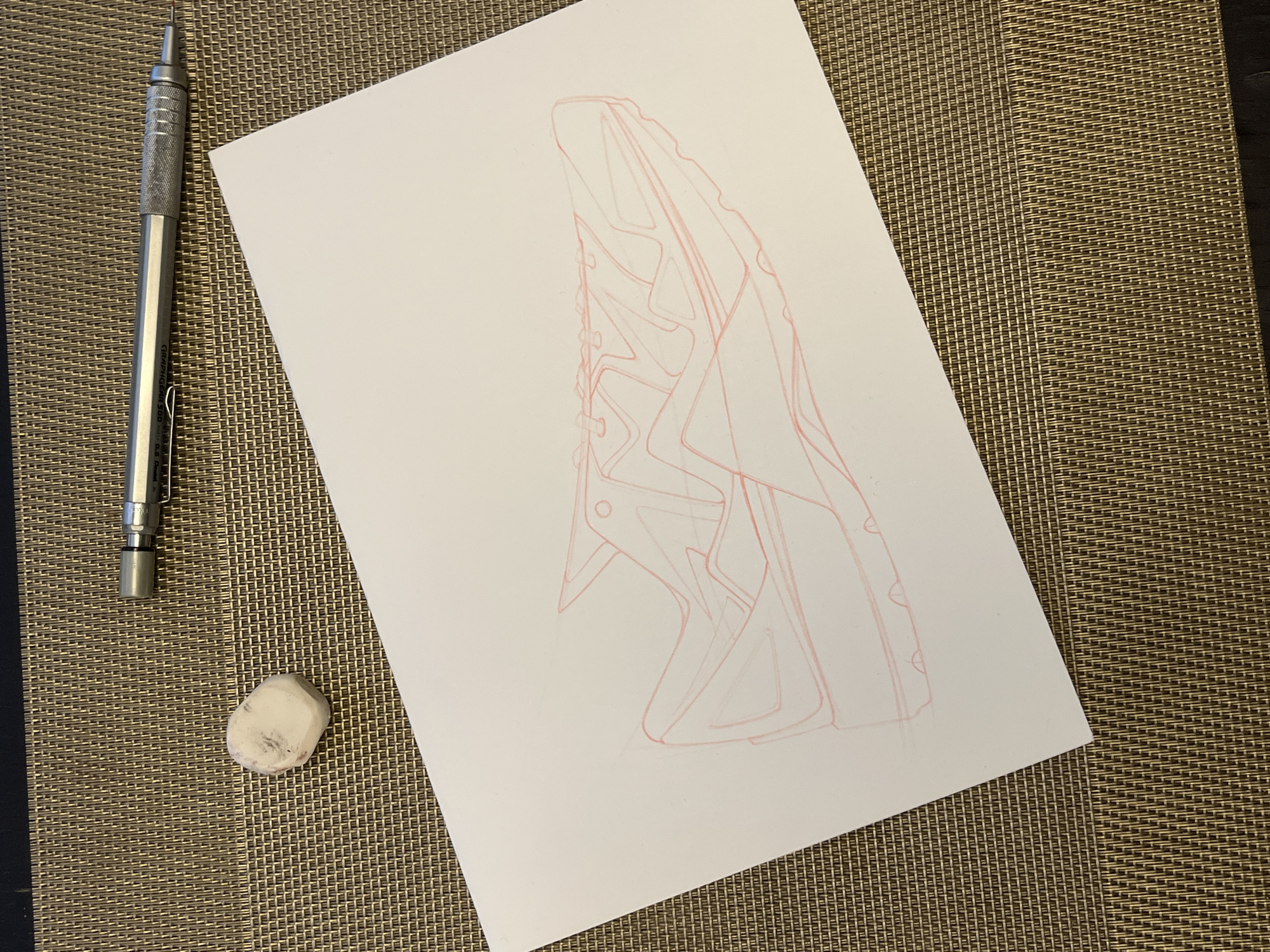 A white sheet of paper with a pencil sketch of a shoe is placed on a brown woven placemat. To the left of the paper are a mechanical pencil and a used white eraser.