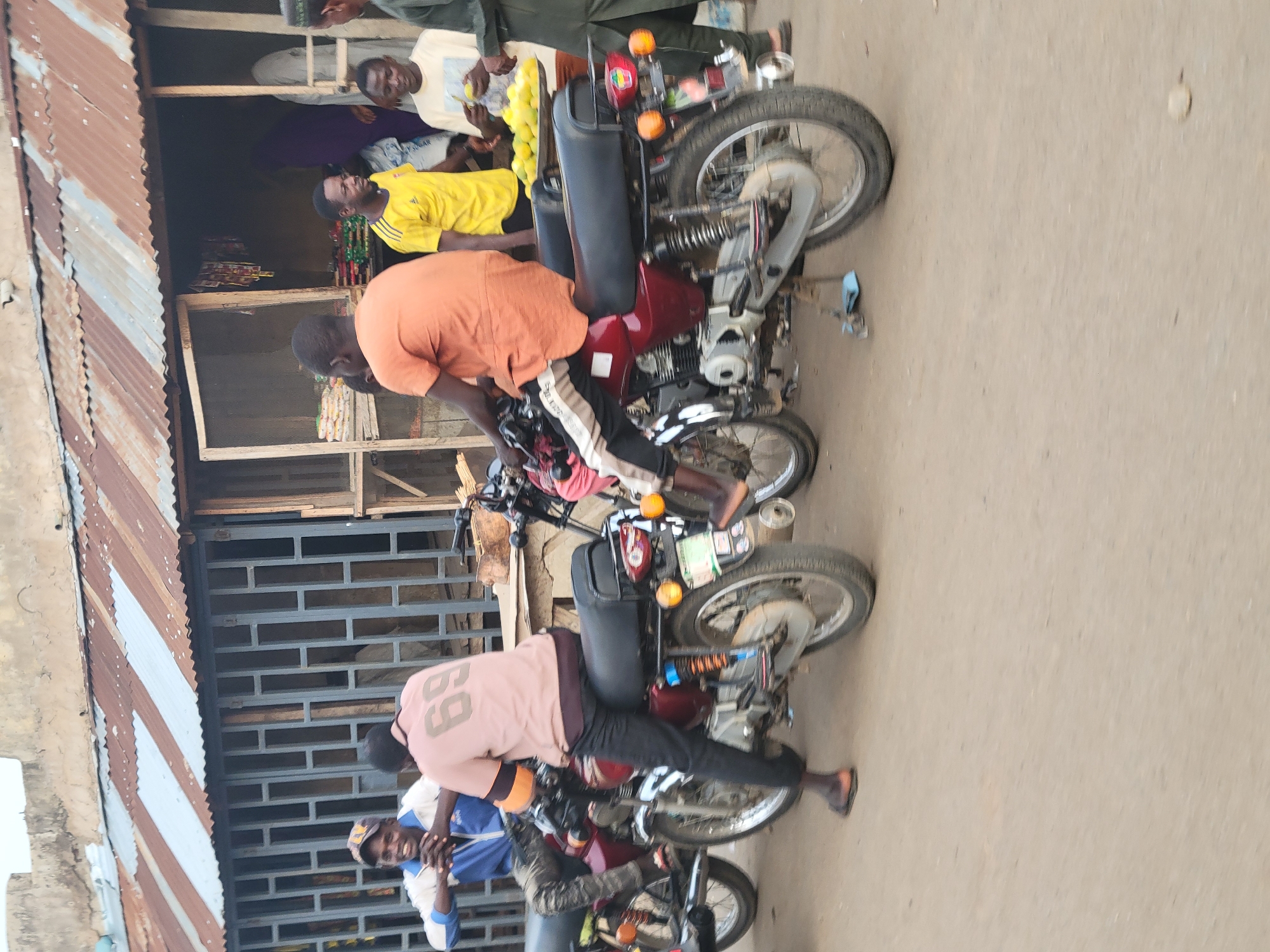 The image shows a street scene in Madalla, Nigeria, with several individuals gathered near a building with a corrugated metal roof. Three motorcycles are parked in a row, each with a rider or person leaning on it. One individual wears an orange shirt and black and white striped pants. Another person is wearing a pink shirt with the number "69" on the back. People are observed standing in the doorway of a building. The ground appears to be dirt or pavement.