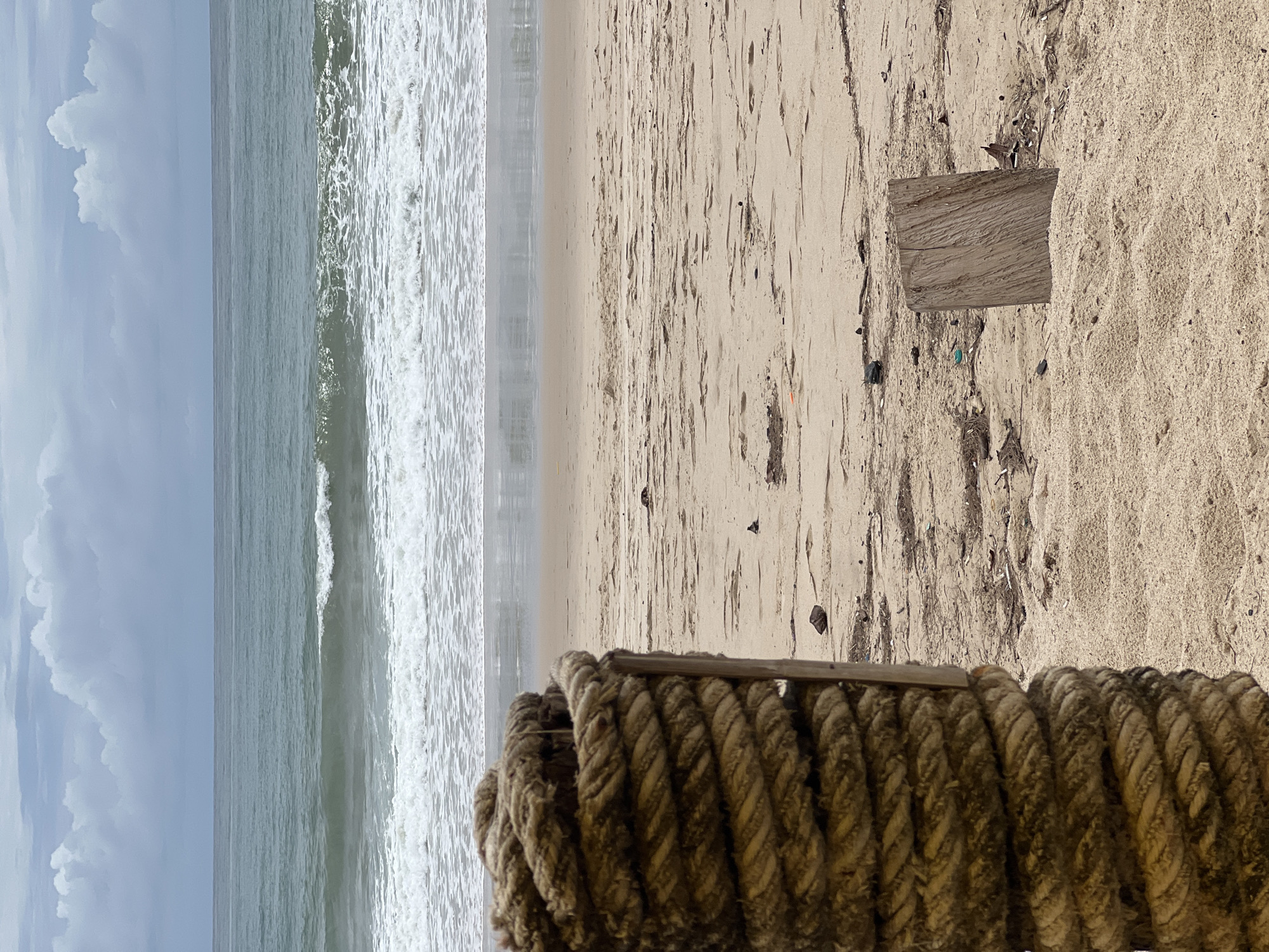 The image shows a beach scene with the ocean on the left and sand on the right. Waves are crashing on the shore. In the foreground, there is a thick rope coiled up, resting on the sand. A rectangular piece of wood is visible further up the beach, partially embedded in the sand. The sky is overcast with clouds. The location is Unknown, Unknown.