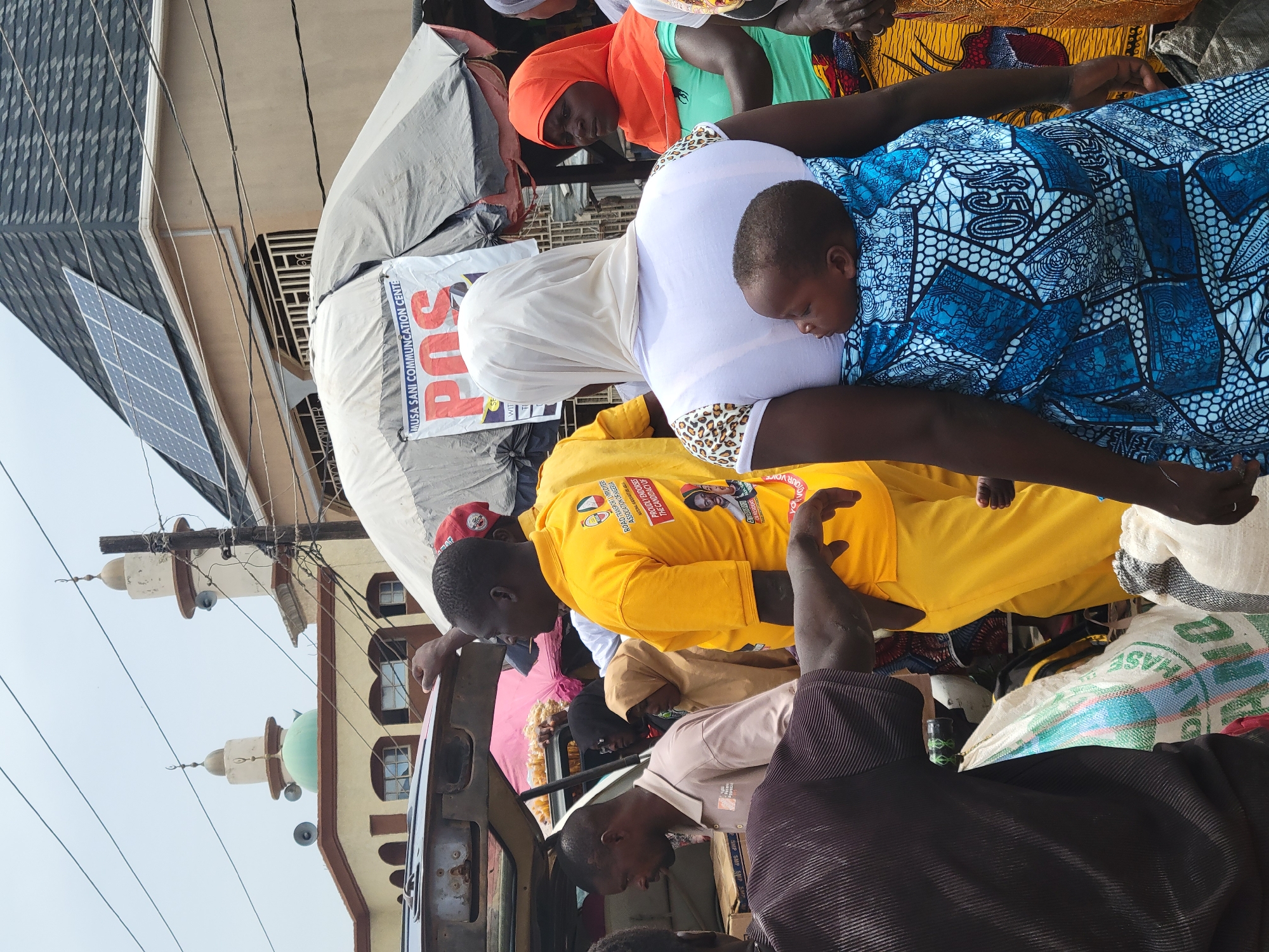 Here's a description of the image:
A diverse scene appears to be taking place in Suleja, Nigeria. A group of people are gathered, some possibly boarding a vehicle.
There is a person wearing a yellow shirt displaying political endorsements, the Road Transport Employers Association of Nigeria, and the Musa Sani Communication Centre. The backdrop features a building with a minaret and solar panels, suggesting the presence of a mosque and modern infrastructure. The environment appears busy, reflecting the vibrancy of the location. There are women in hijabs, possibly traveling with children.
