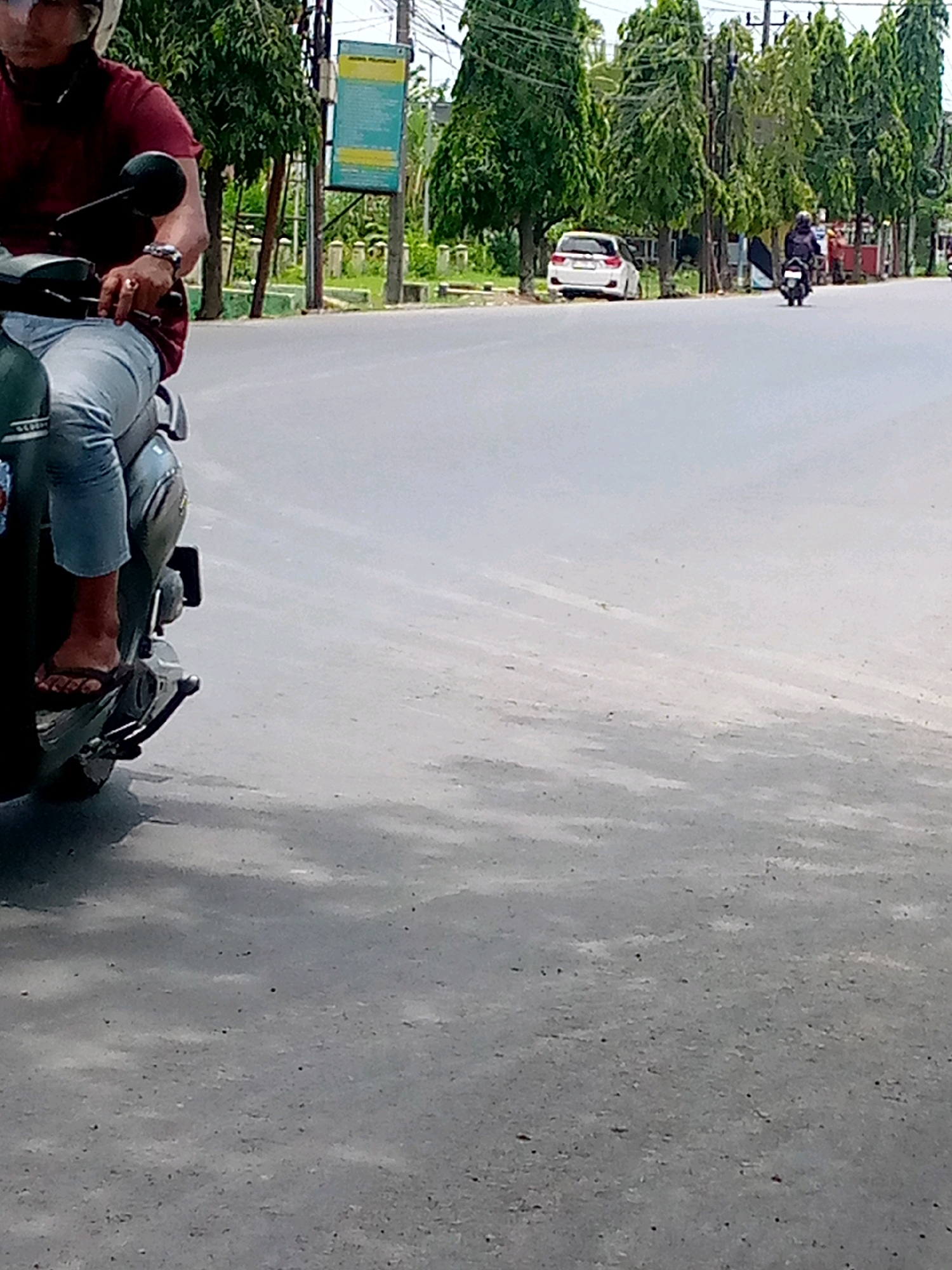 A person wearing a helmet and a maroon shirt is riding a dark-colored scooter. They are wearing light-colored pants and dark-colored flip-flops. The scooter has a dark green body and a silver accent. The person's left hand is on the handlebars, and their right hand appears to be resting on the scooter. In the background, there are trees lining a road, and a white car is parked on the side. Another person on a scooter is further down the road. The scene is set in Keudebayu, Indonesia, on a paved road with some debris.
