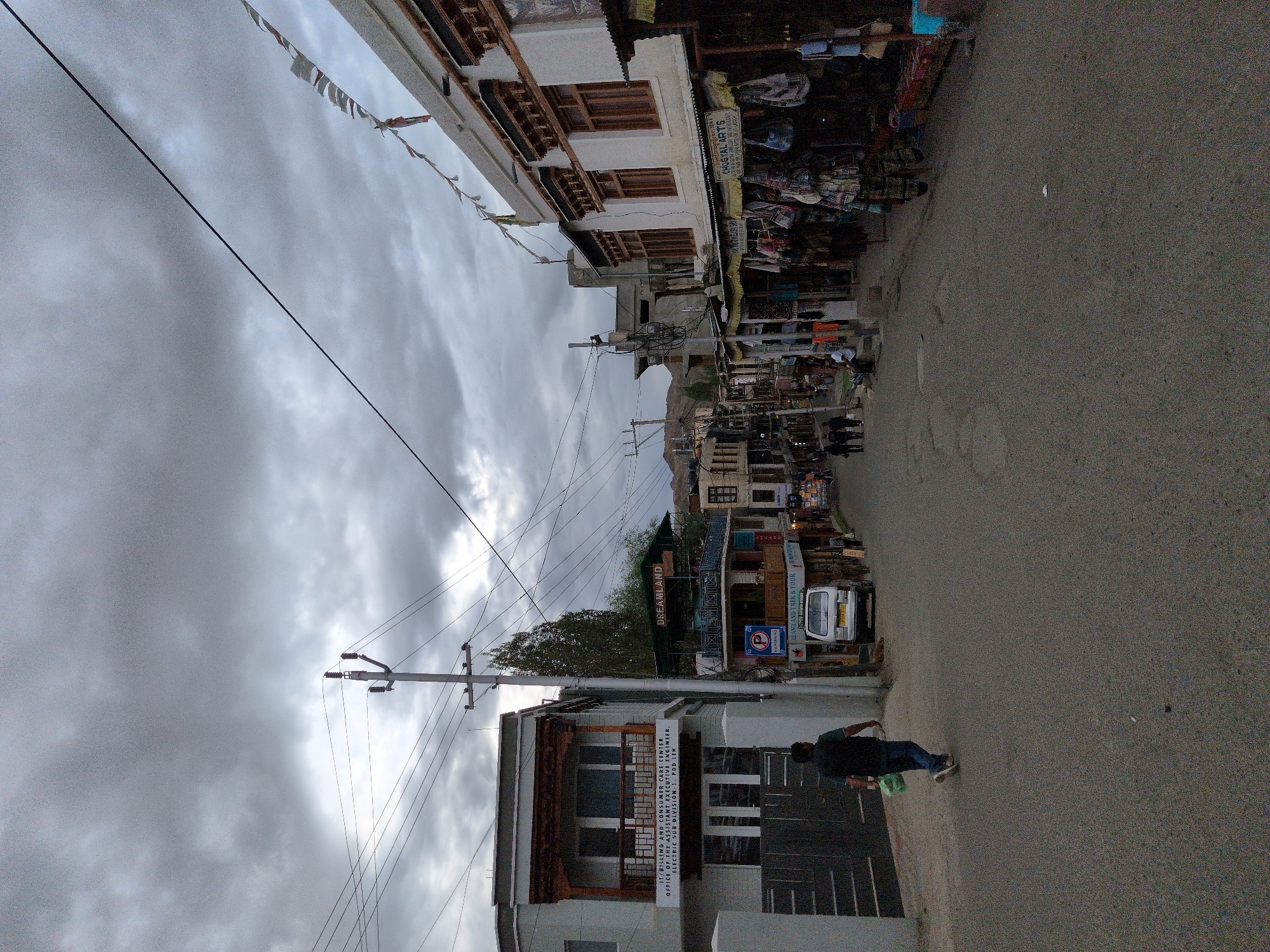 This is a photograph of a street in the city of Leh, India, taken on a cloudy day. The street is paved with asphalt and is lined with buildings on both sides. The buildings are a mix of traditional and modern architecture, with some featuring intricate wooden carvings. On the left side of the street, there is a multi-story building with a balcony and several windows. A sign on this building reads "IT/BILLING AND CONSUMER CARE CENTER OFFICE OF THE ASSISTANT EXECUTIVE ENGINEER, ELECTRIC SUB DIVISION-1, PDD". A person in a dark vest and blue jeans is walking away from the camera on the left side of the street, carrying a light green bag.
On the right side of the street, there are several shops with awnings displaying various goods, likely souvenirs or textiles. Signs above the shops read "DREAMLAND" and "CHOGYAL ARTS". A white van is parked on the right side of the street near the shops. In the distance, more buildings and hills are visible under the overcast sky. Prayer flags are strung across some of the buildings, adding a touch of color to the scene. The overall impression is of a busy commercial street in a Himalayan town.