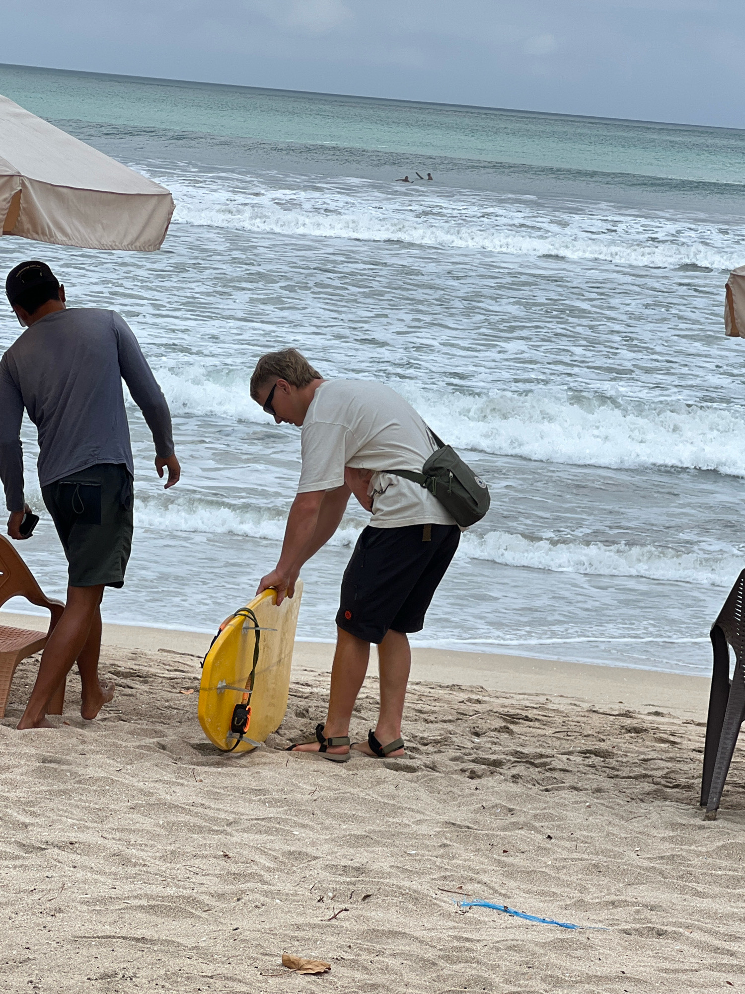 Two men are on a sandy beach in Kuta, Indonesia, near the ocean. The man on the left, wearing a grey long-sleeved shirt and dark green shorts, is holding a black smartphone in his right hand. He is facing the ocean. The man on the right, wearing a light-colored short-sleeved shirt and black shorts, is bending over and holding a yellow surfboard with orange and black straps. He is also facing the ocean, and has a small green bag slung across his chest. Waves are breaking on the shore in the background, and the sky is overcast. A large beach umbrella is visible on the left side of the image, and a dark chair is partially visible on the right.