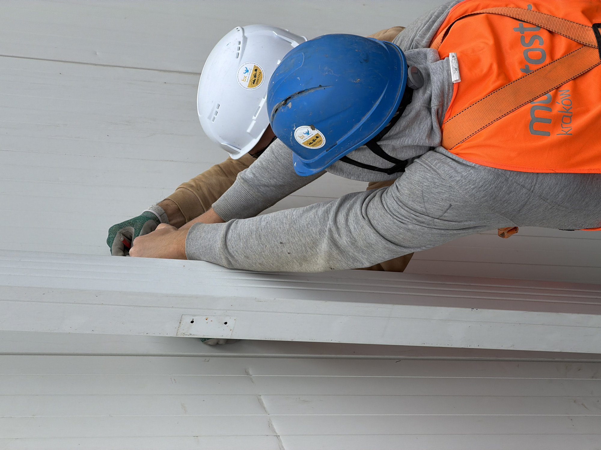 Two construction workers in hard hats and safety vests are seen from the chest up, working on a white corrugated wall in Stróża, Poland. The worker in the foreground wears a blue hard hat and an orange safety vest with "motostart kraków" printed on it. The worker behind him wears a white hard hat. Both are wearing gloves. The worker in the foreground is holding a piece of white corrugated material, appearing to install it into the wall. The background is a plain white wall with vertical corrugated lines.