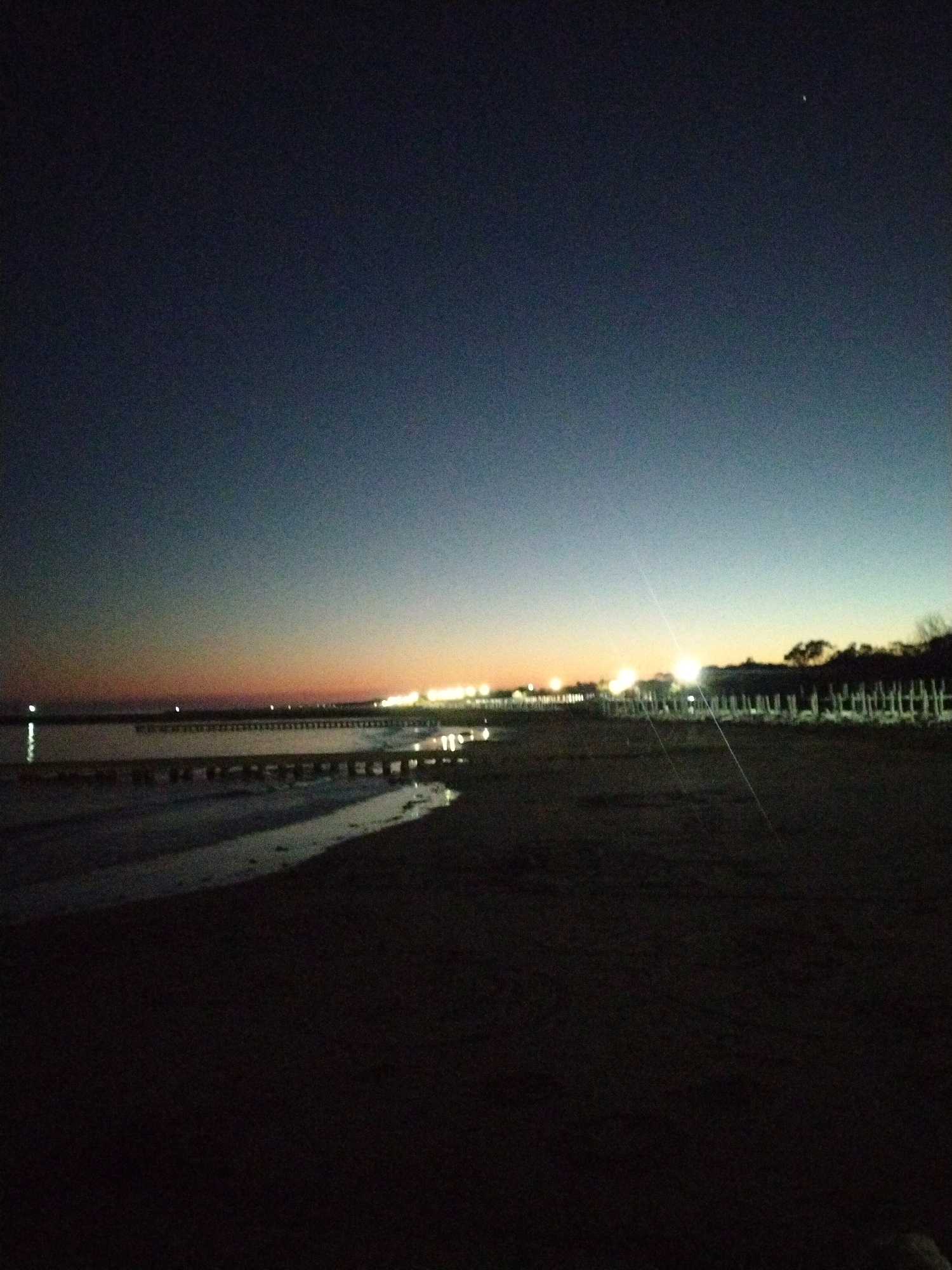 This image captures a beach scene at twilight in Caorle, Italy. The sky displays a gradient of dark blue at the top, transitioning to lighter blues and then to oranges and pinks near the horizon. A faint star is visible in the upper right portion of the sky.
In the middle ground, a body of water, likely the Adriatic Sea, is present. A wooden pier or structure extends horizontally across the water, with several lights illuminating its surface. The water reflects these lights, creating streaks of light. Another, closer structure, also appears to be a pier or walkway, runs parallel to the shoreline.
The foreground consists of a sandy beach, largely in shadow. A narrow strip of water, possibly a tidal pool or a shallow inlet, cuts through the sand, reflecting a faint light.
In the distance, along the horizon, a line of trees and buildings are silhouetted against the fading light, with numerous bright lights indicating a populated area. The overall scene conveys a sense of calm and quiet, characteristic of a beach at the end of the day.