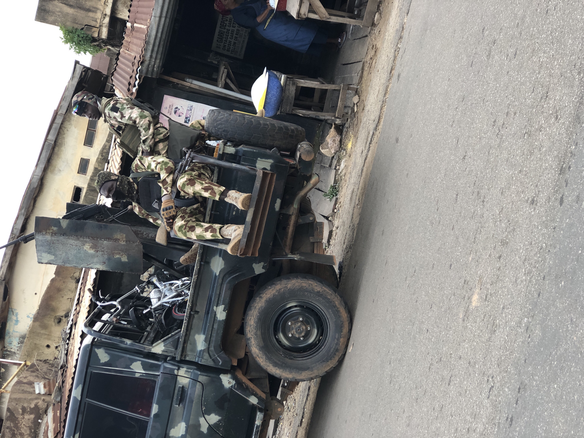 In Jos, Nigeria, two soldiers in camouflage uniforms are seated in the back of a military vehicle, a dark-colored pickup truck with a camouflage pattern. The truck is loaded with what appears to be a small bicycle and other equipment. The soldier in the foreground is wearing a patterned head covering and a tactical vest, with a rifle resting across his lap. The soldier behind him is wearing a helmet with goggles and also has a tactical vest. They are positioned on the edge of the truck bed, overlooking a street scene.
Adjacent to the truck is a roadside stall with a wooden structure and a corrugated metal roof. On the stall, a large blue bowl filled with a white substance, possibly food, is placed on a wooden stool. A woman in a blue apron and headscarf is standing at the stall, appearing to be working or serving customers. A sign on the stall reads "M.G GARRI DEPOT" followed by further text.
The environment suggests a bustling urban or semi-urban setting with older buildings visible in the background, some with damaged walls and roofs. The road in the foreground is paved and shows signs of wear. The overall impression is of a potentially active or vigilant presence in a commercial area of Jos.