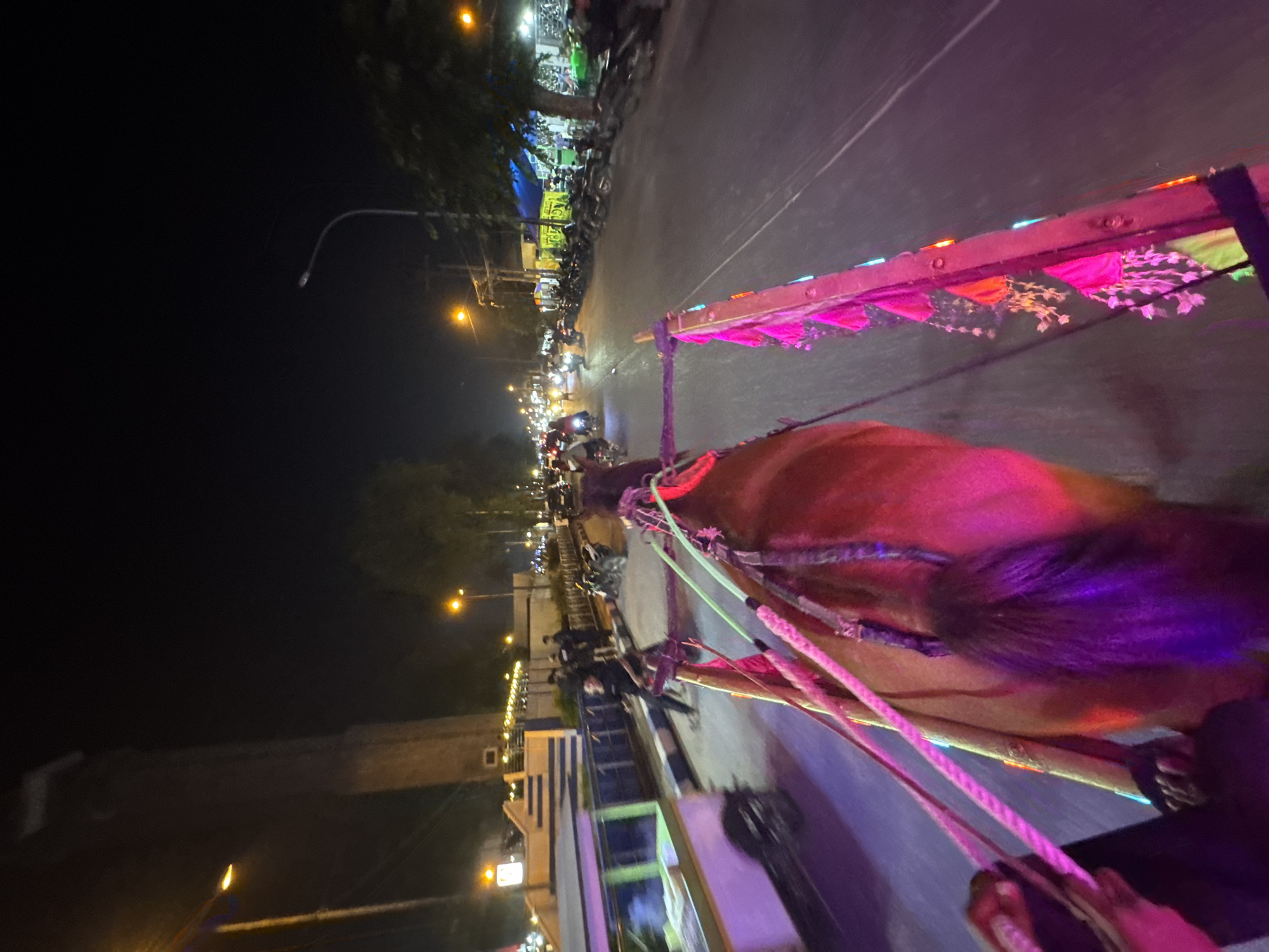 The image captures a nighttime scene in Lubuk Pakam, Indonesia, from the perspective of a moving horse-drawn carriage. The carriage itself is adorned with bright pink and blue lights, and colorful tassels hang from its sides. A horse, partially visible in the foreground, pulls the carriage. The road is paved, and traffic, including motorcycles and cars, is present. Streetlights illuminate the scene, and buildings line the roadside. In the background, a tall, dark structure, possibly a tower, is visible, illuminated by yellow lights. The overall atmosphere is lively, suggesting a public event or a busy night in the city.
