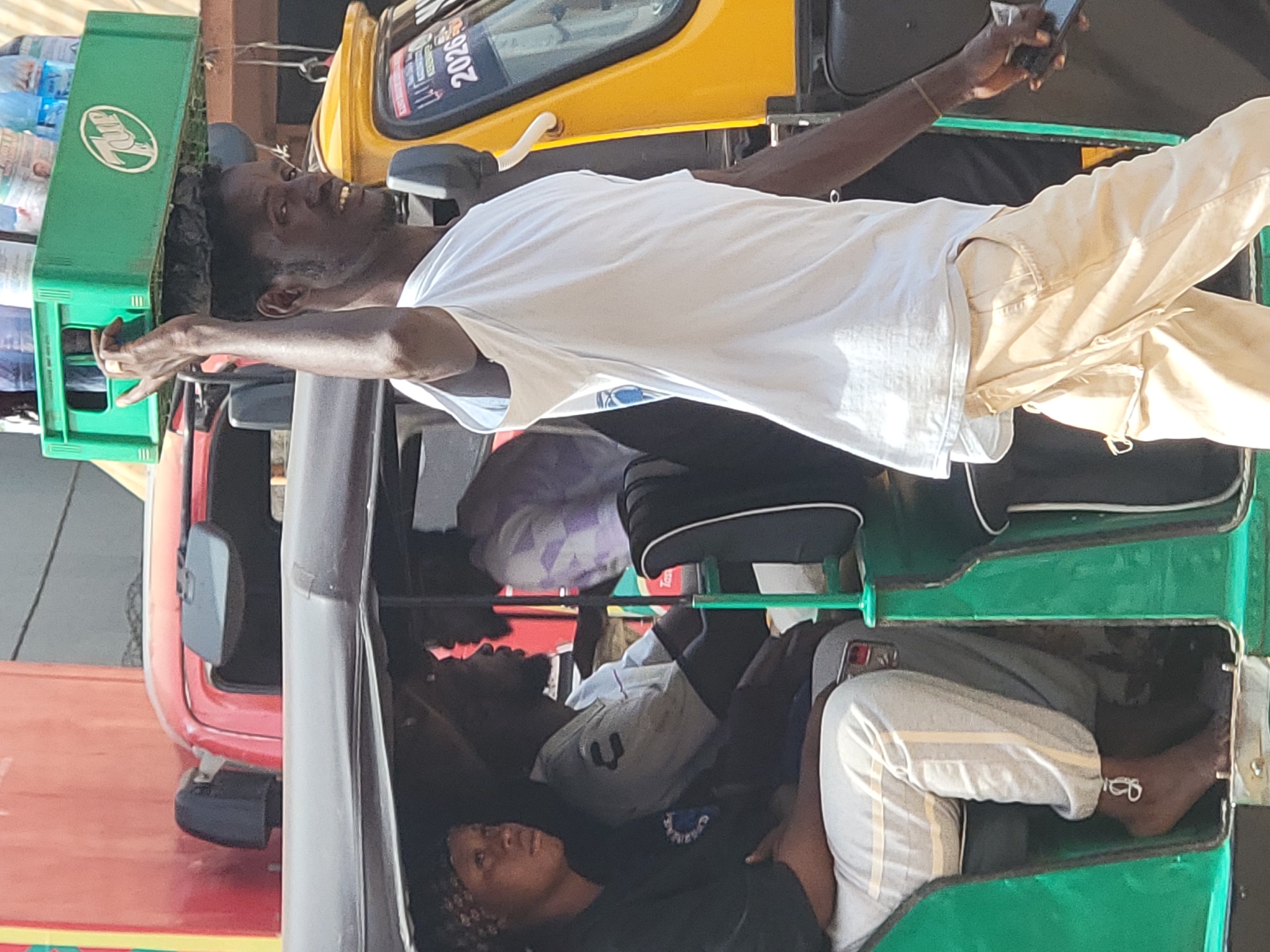 In Maiburiji, Nigeria, several individuals are tightly packed inside a three-wheeled vehicle. A man wearing a white t-shirt and khaki pants is leaning out of the vehicle's open top, holding a mobile phone. A crate of 7-Up bottles sits on top of the vehicle. Other individuals are visible inside the vehicle's passenger compartment. A red vehicle is partially visible to the left. A yellow vehicle is partially visible above and to the right.