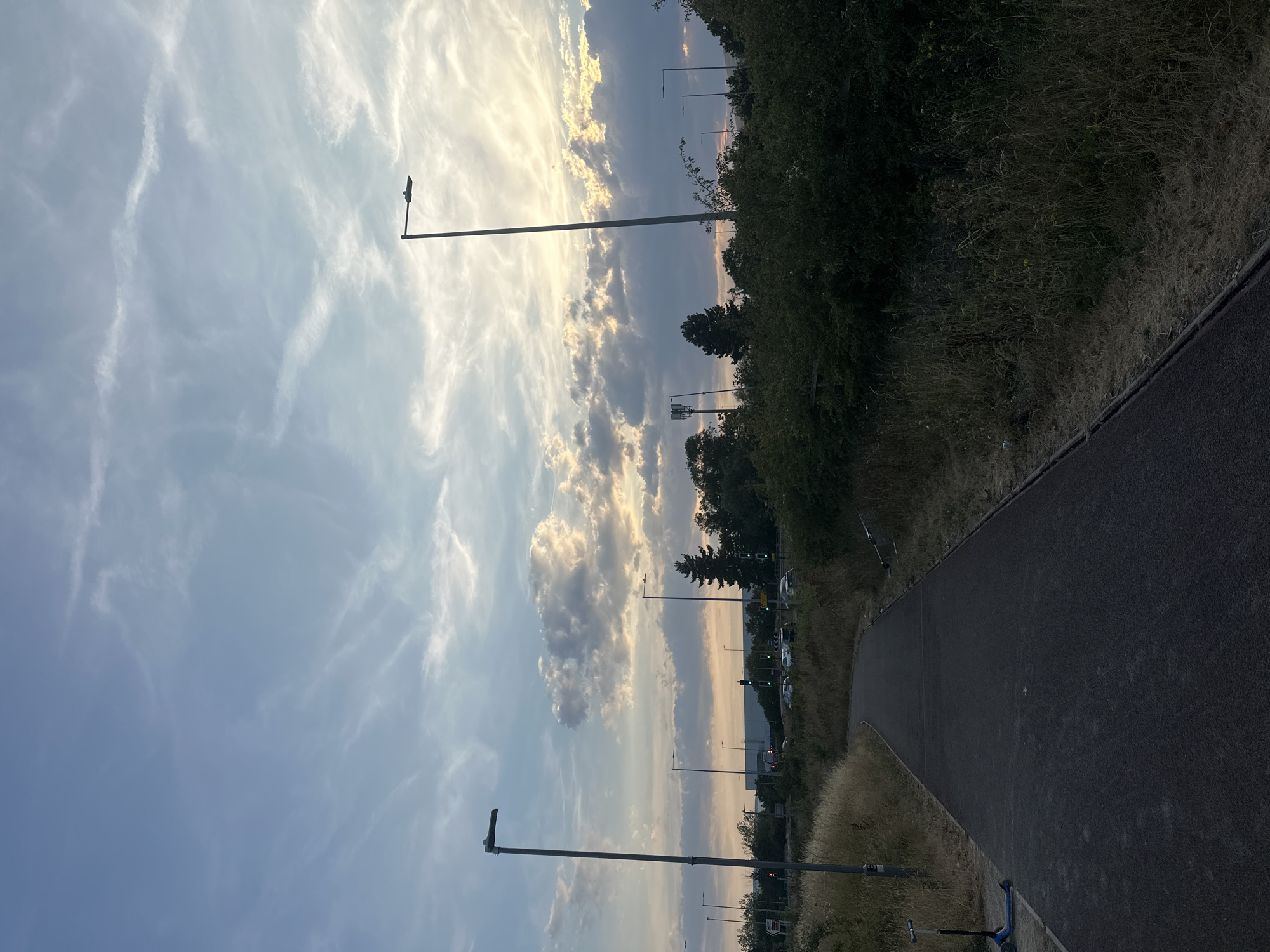 This is an outdoor, eye-level, full shot of a paved pathway in Milton Keynes, United Kingdom, at dusk. The pathway, which appears to be a pedestrian or cycle path, curves to the left and is bordered by dry grass and bushes on either side. In the foreground, a blue and black scooter is parked on the left side of the path. The sky above is filled with clouds, with the setting sun casting a warm glow through them. Several tall streetlights are visible throughout the scene, with one prominently positioned to the left of the path. In the background, trees and buildings are silhouetted against the sky, and traffic lights and cars can be seen on a distant road. The overall atmosphere is peaceful and serene, capturing the quiet end of the day in a suburban or semi-rural setting.