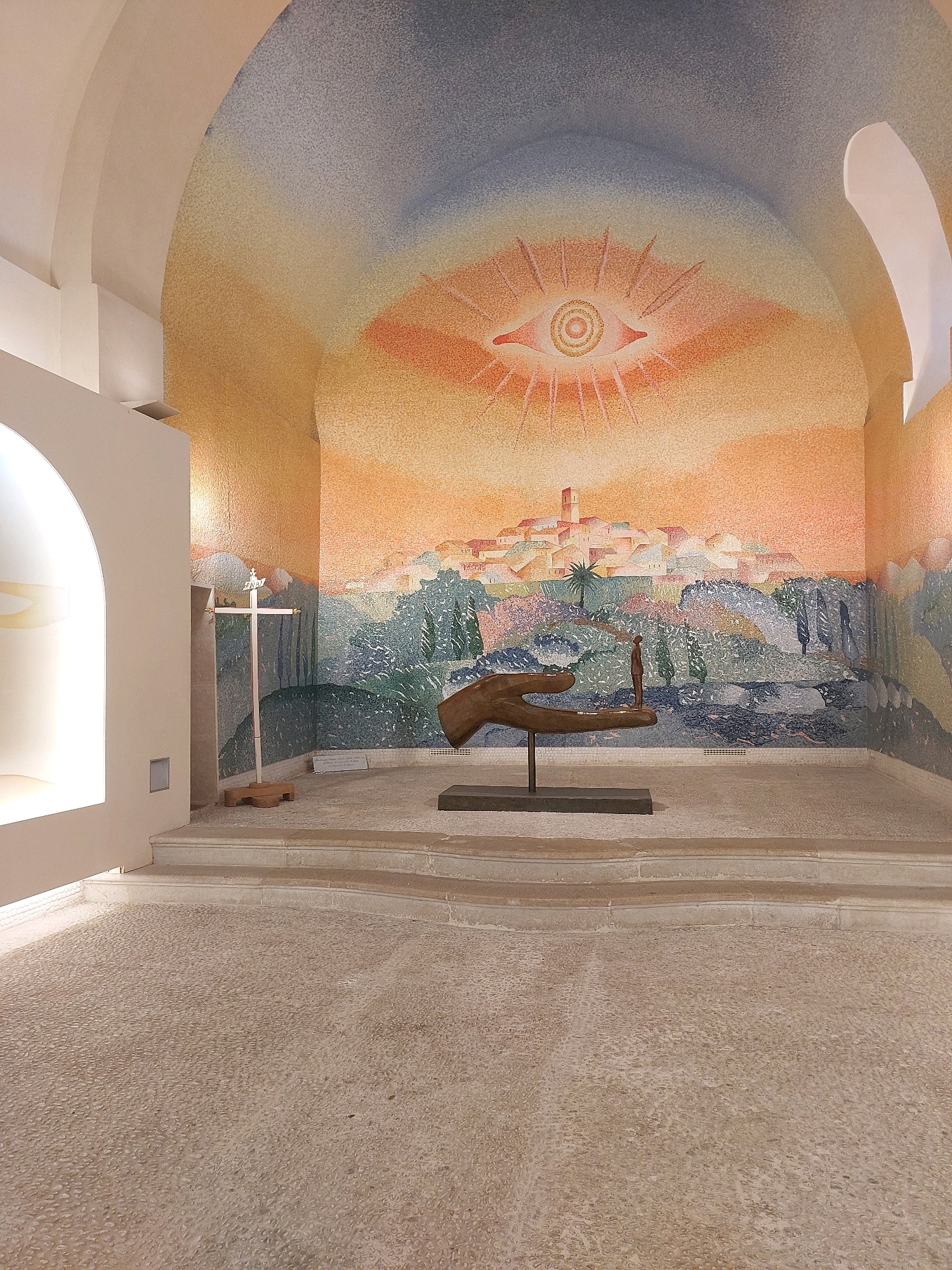 The image depicts the interior of the Chapelle du Rosaire, also known as the Matisse Chapel, located in Vence, France, near Saint-Paul-de-Vence. The chapel features a large mosaic mural that covers the curved walls and ceiling. The mural depicts a stylized landscape with a village, trees, and a prominent eye symbol at the apex of the ceiling, radiating rays. In the foreground, a bronze sculpture of an open hand holding a small human figure stands on a dark rectangular base. To the left of the sculpture, a wooden cross with a white crucifix stands on a wooden base, situated on a raised platform. The floor is covered in small, light-colored pebbles. The architecture of the chapel includes curved ceilings and white walls.