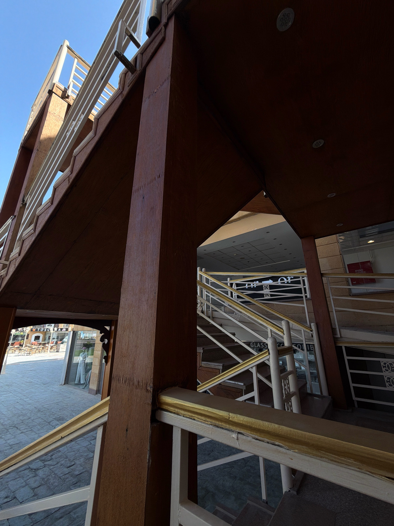 The image captures a low-angle view of an outdoor staircase and walkways constructed with wooden beams and white railings accented with gold trim. The architecture suggests a building or an elevated section of a complex, possibly a commercial area or resort. The background reveals a clear blue sky. Beneath the elevated walkway, an outdoor seating area with tables and chairs is visible, along with shop windows displaying merchandise. One shop has a sign that reads "TOSCANA". The flooring in the foreground and in the open area appears to be paved with hexagonal tiles. The overall scene is set in a sunny, urban environment.