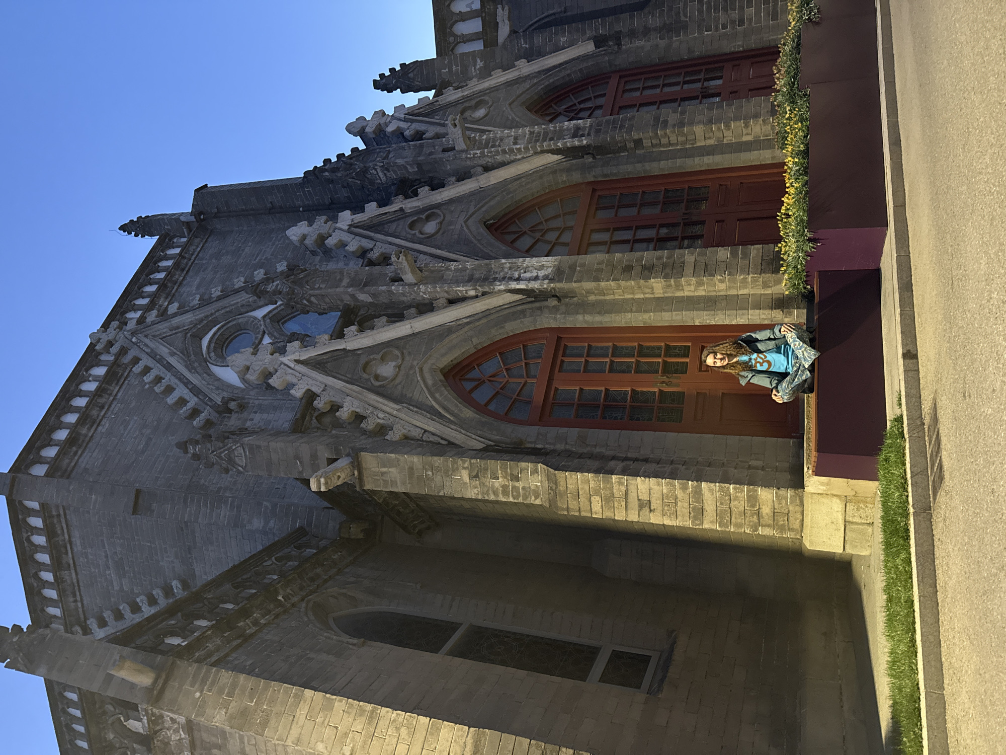 A low-angle, wide shot depicts the side of a stone building, adjacent to a sidewalk, on which a person is seated. The person is a woman with long hair. She is wearing a blue t-shirt, a gray jacket, and blue patterned pants. She is sitting on a structure made of gray bricks with a painted dark-red top. The building is constructed of gray bricks, with multiple Gothic architectural elements, including windows with pointed arches, decorative stone ornamentation, and a gabled roof line. The windows have red frames. A patch of greenery is situated between the building and the sidewalk. The sky is blue. The image appears to be taken at dusk or dawn. It is in Xicheng District, China.