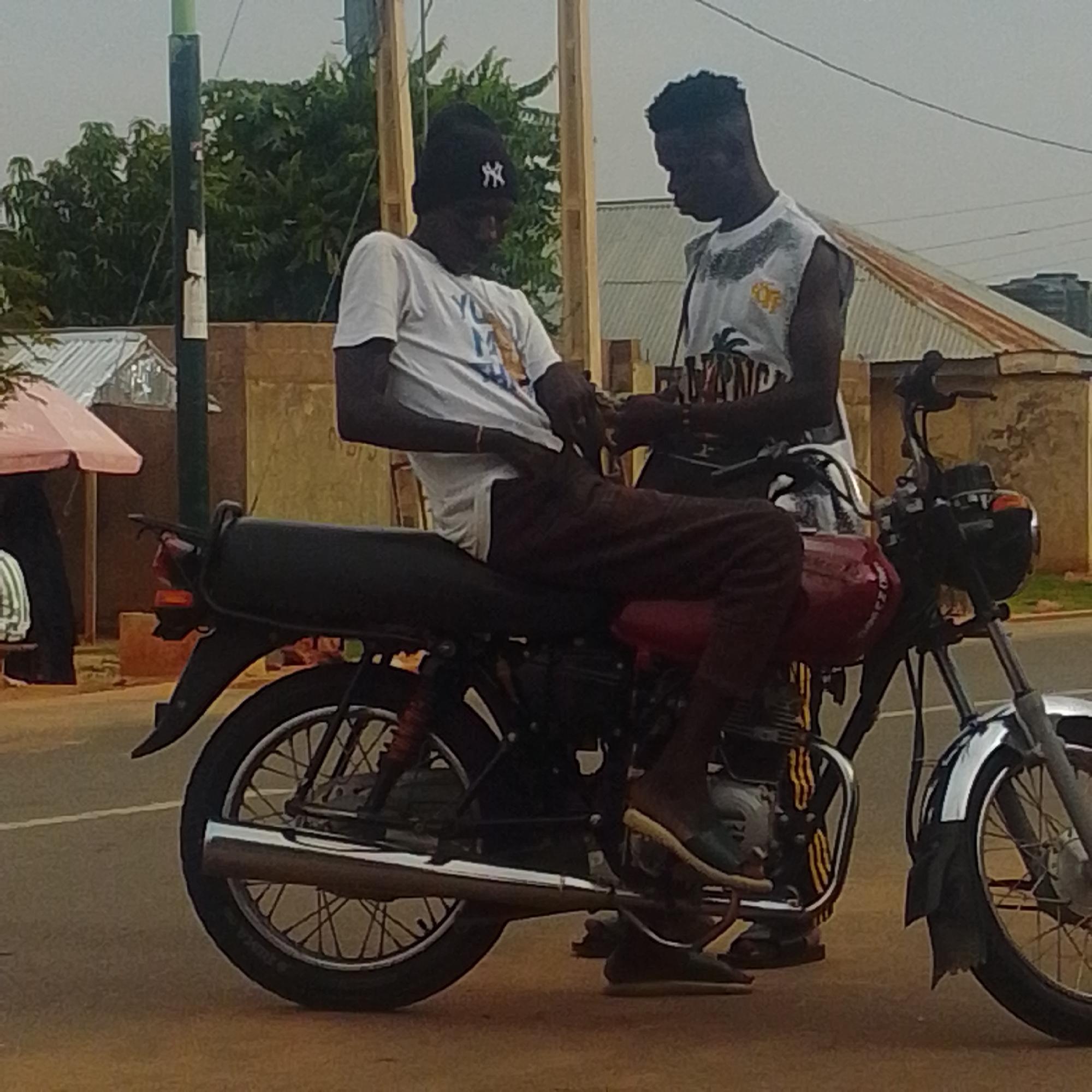 Two young Black men are on a motorcycle, which is stopped on a paved road. The man in the front is wearing a white t-shirt and a black beanie with a white logo. He is sitting on the motorcycle, and the man behind him is standing, leaning forward and appearing to adjust something on the front of the motorcycle. The man behind him is wearing a white sleeveless shirt with a grey and black pattern around the neckline and what looks like palm trees and text printed on it. The motorcycle is red and black, with a silver exhaust pipe and silver handlebars.
In the background, there are trees, buildings with corrugated metal roofs, and a utility pole. The sky is overcast, suggesting it is daytime, possibly early morning or late afternoon. The overall scene appears to be in a semi-urban or rural area in Gombe, Nigeria. The men's interactions suggest a moment of repair or preparation before continuing their journey. The image has a somewhat grainy quality, indicating it was taken with a mobile phone camera or in low light conditions.