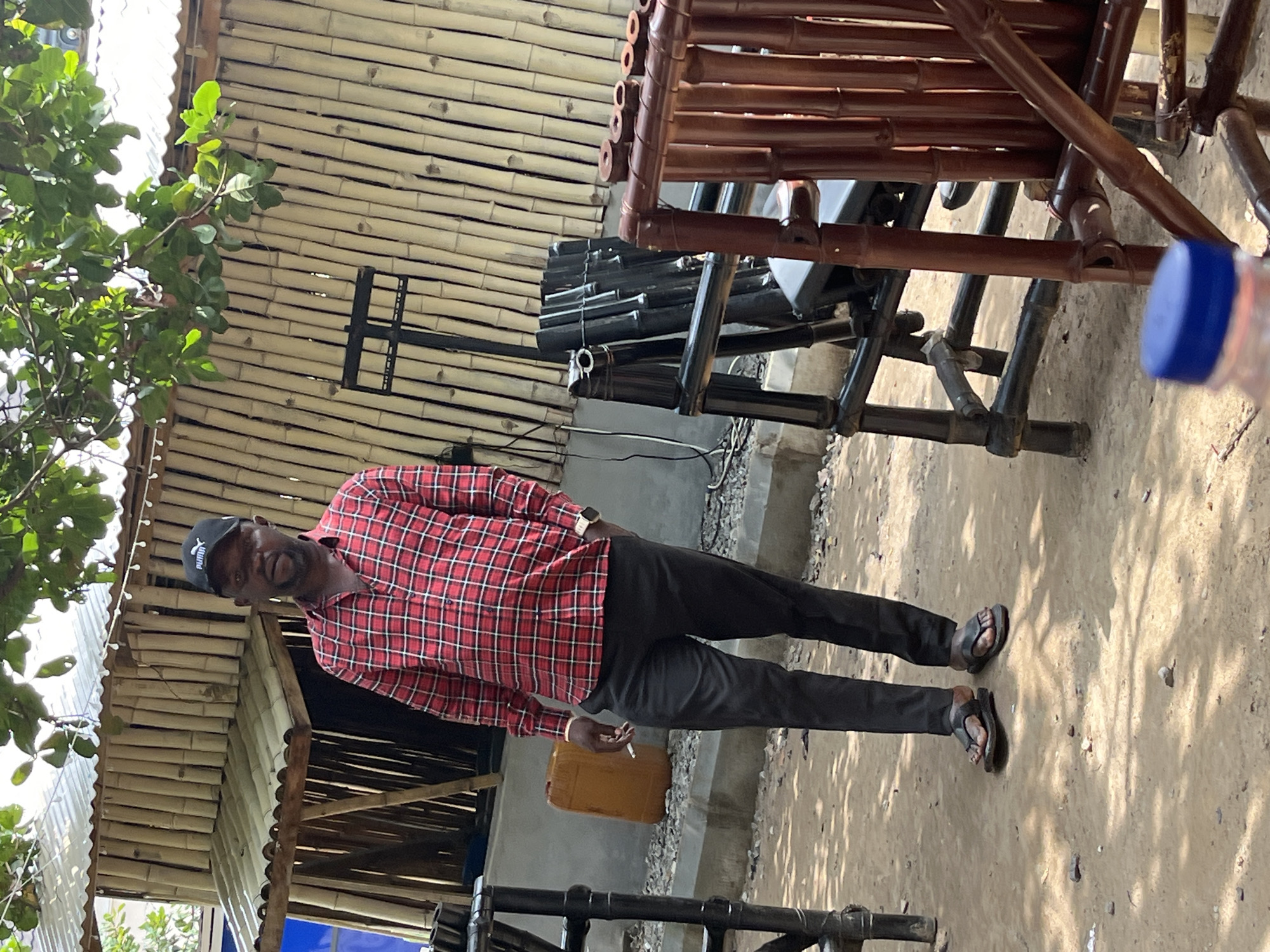 A man wearing a red and black plaid shirt, black pants, a black baseball cap, and dark flip-flops stands in an outdoor setting. He is holding a cigarette in his right hand and appears to be looking towards the camera with a slight smile. Behind him, there is a structure made of bamboo poles and a wooden table. The ground is covered with dirt and gravel. In the background, there are more bamboo structures and some greenery. A blue-capped bottle is visible on the right side of the image. The lighting suggests it is daytime, possibly sunny, with shadows cast on the ground. The scene appears casual and possibly rural or semi-urban. There is no visible text in the image, except for a logo on the man's cap which appears to be "Puma".