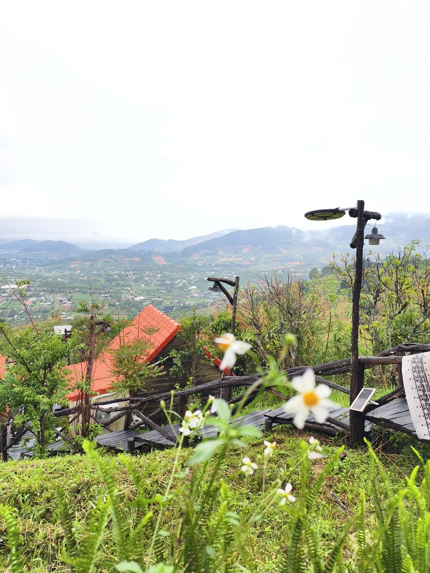 The image captures a scenic view from an elevated position, likely in a mountainous or hilly region. In the background, there's a sprawling landscape with hints of a city or town amidst the green valleys and distant mountains, shrouded in a light mist.
In the foreground, there's a wooden pathway or bridge, suggesting a tourist spot or a resort. A rustic wooden light post stands on the side, equipped with what seems to be a solar panel. Closer to the camera, there are vibrant green plants, including ferns and small white flowers with yellow centers, adding a touch of natural beauty to the constructed landscape.
The overall atmosphere is serene and peaceful, emphasizing the blend of nature and human construction.