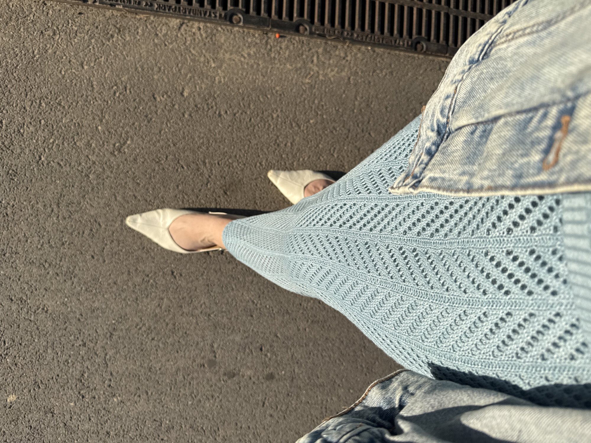 An overhead shot shows a person standing on a paved surface, likely a sidewalk or street. They are wearing a light blue, knitted dress with a chevron pattern and a denim jacket. White, pointed-toe shoes are visible on their feet. To the right of the person, a metal grate or drain cover is embedded in the pavement, with the words "STANDARD PARK" visible on its surface. The pavement itself appears worn and textured, with shadows cast by the sun indicating it is daytime. The setting does not provide specific clues to its location within Ufa, Russia.