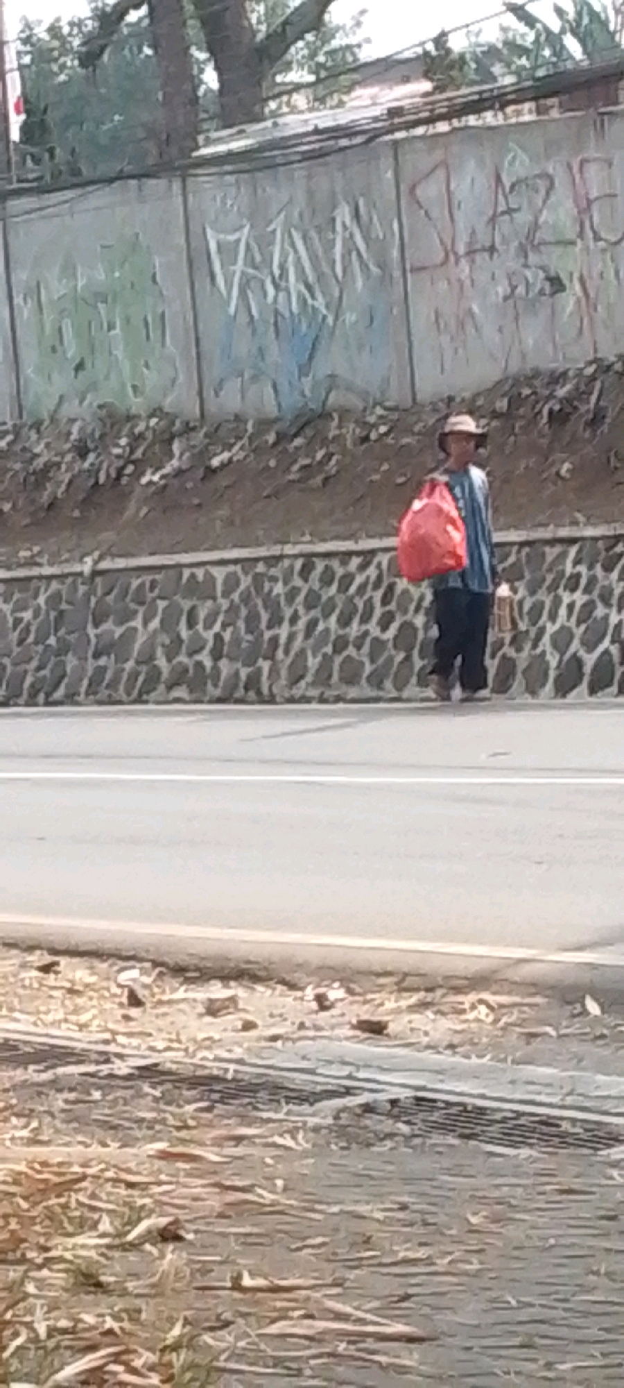A person wearing a hat and a blue and white striped shirt walks along the side of a road. They are carrying a large red bag over their left shoulder and a brown bottle in their right hand. The person is walking on the shoulder of a paved road with a white line marking the lane. Behind the road is a textured stone wall, and above that, a slope covered with dirt and rocks, leading up to a concrete wall covered in graffiti. Trees and foliage are visible above the graffiti wall. The scene appears to be in an outdoor urban or semi-urban environment, possibly near Cilaku, Indonesia, given the context.